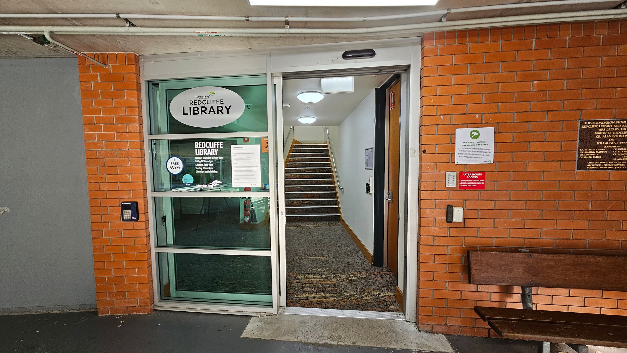 A brick wall with a glass door on the left, displaying "Redcliffe Library" signage and information about free Wi-Fi. The door is open, revealing a carpeted staircase leading upwards. On the right, there are plaques and signs on the wall, including one about after-hours access. A wooden bench is positioned against the wall. The ceiling has exposed pipes and lights, and the floor is concrete. A brick wall with a glass door on the left, displaying "Redcliffe Library" signage and information about free Wi-Fi. The door is open, revealing a carpeted staircase leading upwards. On the right, there are plaques and signs on the wall, including one about after-hours access. A wooden bench is positioned against the wall. The ceiling has exposed pipes and lights, and the floor is concrete.