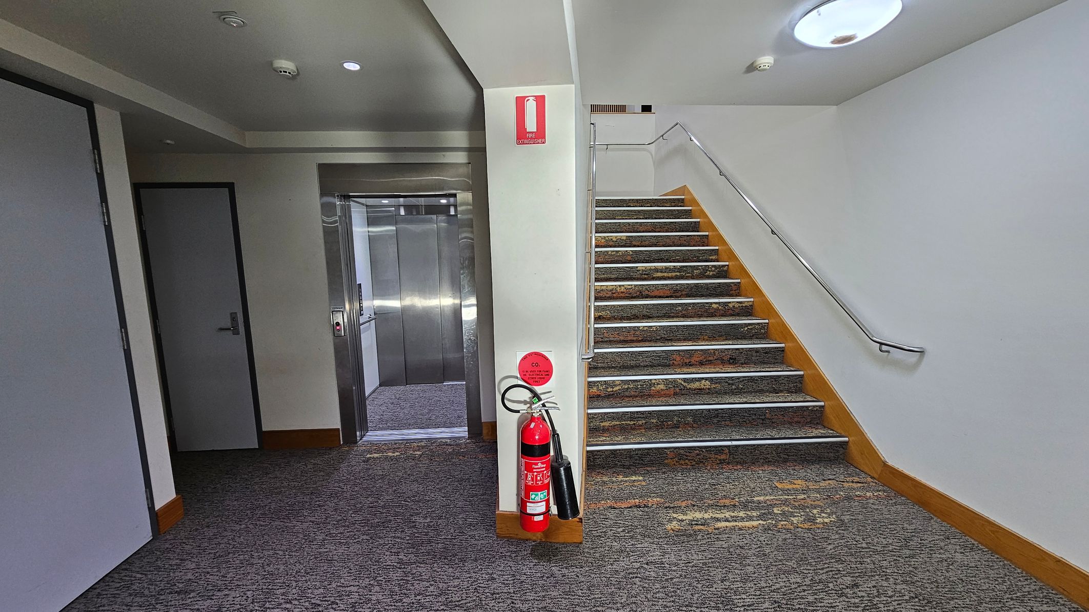 A hallway with a grey carpeted floor. On the left, there is a closed door and an open lift with metallic doors. In the centre, a fire extinguisher is mounted on a white wall. To the right, a staircase with a patterned carpet and a metal handrail ascends. The ceiling is white with recessed lighting and a smoke detector. A hallway with a grey carpeted floor. On the left, there is a closed door and an open lift with metallic doors. In the centre, a fire extinguisher is mounted on a white wall. To the right, a staircase with a patterned carpet and a metal handrail ascends. The ceiling is white with recessed lighting and a smoke detector.