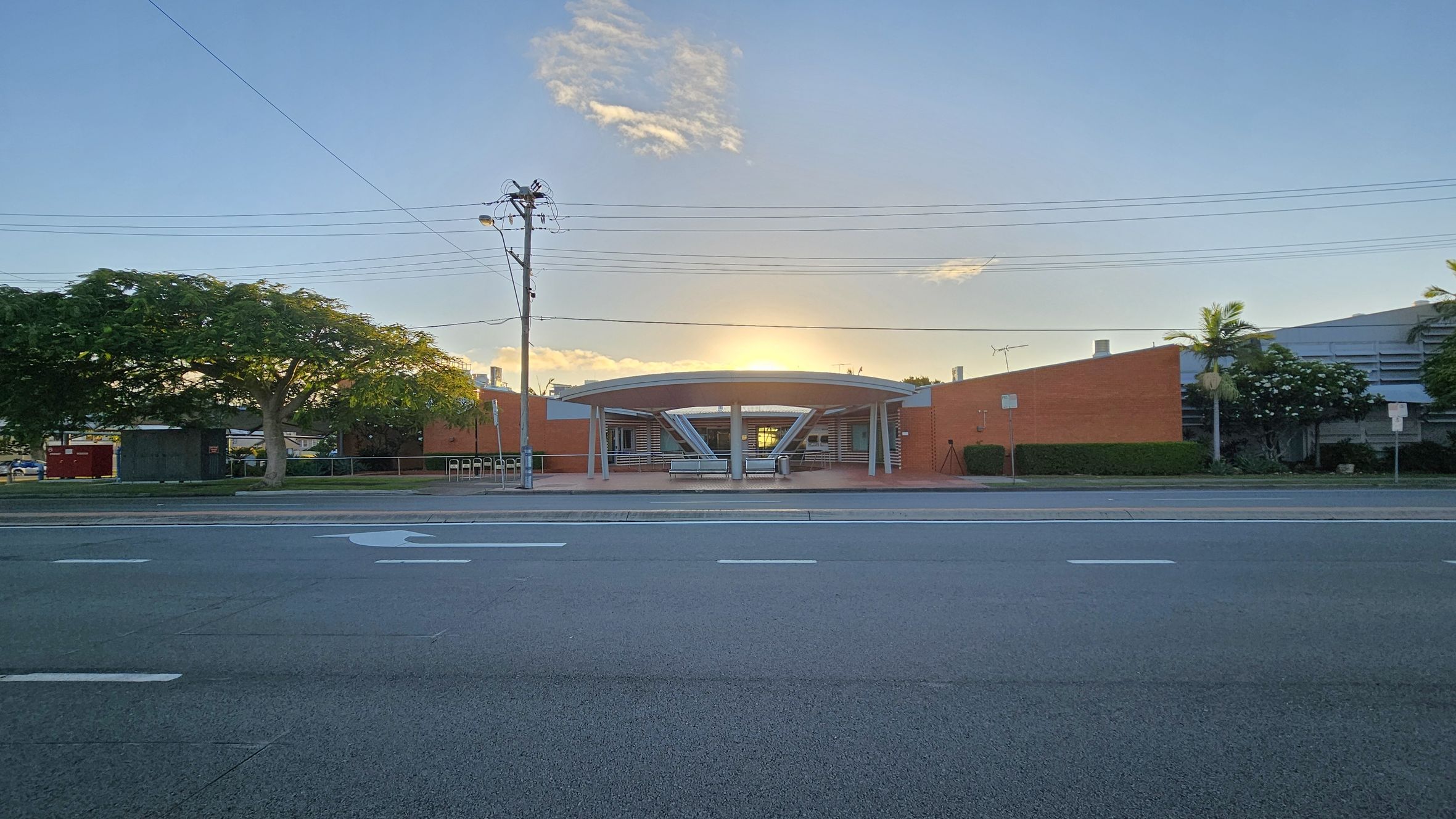 A wide road in the foreground with white lane markings. In the middle, a building with a circular entrance and a flat roof, flanked by red brick walls. A large tree with a thick trunk and green foliage is on the left, while smaller trees and shrubs are on the right. Overhead, power lines stretch across the scene. The sun is setting behind the building, casting a warm glow. A wide road in the foreground with white lane markings. In the middle, a building with a circular entrance and a flat roof, flanked by red brick walls. A large tree with a thick trunk and green foliage is on the left, while smaller trees and shrubs are on the right. Overhead, power lines stretch across the scene. The sun is setting behind the building, casting a warm glow.