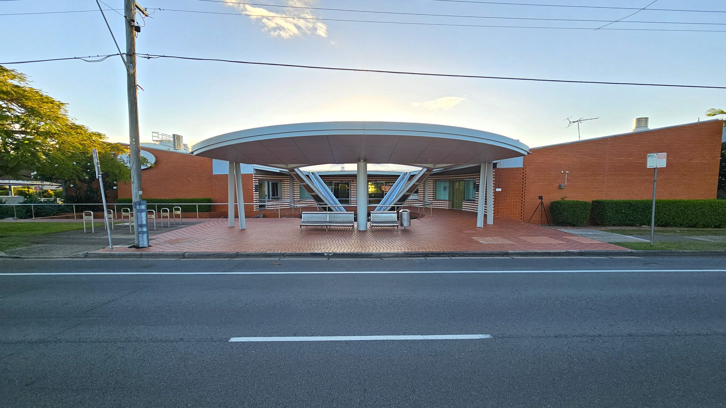 A modern building with a circular entrance featuring a large, round canopy supported by angled metal beams. Two benches are placed symmetrically under the canopy. The structure is made of red brick with horizontal white accents. A road runs in the foreground, and a power pole with wires is on the left. Green hedges and a tree are visible on the left side, while a small signpost is on the right. A modern building with a circular entrance featuring a large, round canopy supported by angled metal beams. Two benches are placed symmetrically under the canopy. The structure is made of red brick with horizontal white accents. A road runs in the foreground, and a power pole with wires is on the left. Green hedges and a tree are visible on the left side, while a small signpost is on the right.