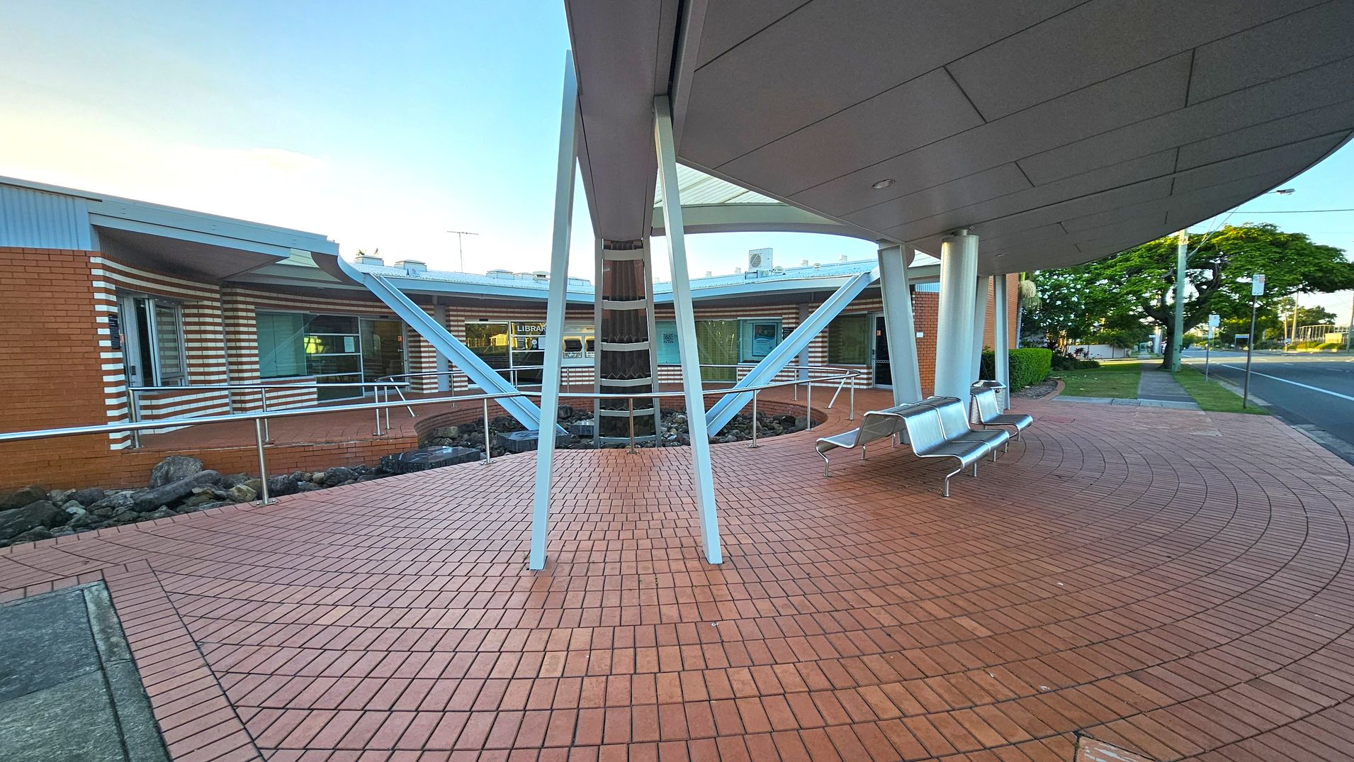 A modern building with a curved design featuring red brick walls and white horizontal stripes. The foreground has a tiled area with metal benches. The structure is supported by angled metal beams. A glass window displays the word "LIBRARY" inside. To the right, a road runs alongside a grassy verge with trees and a lamppost. The ceiling is smooth and white, with recessed lighting. A modern building with a curved design featuring red brick walls and white horizontal stripes. The foreground has a tiled area with metal benches. The structure is supported by angled metal beams. A glass window displays the word "LIBRARY" inside. To the right, a road runs alongside a grassy verge with trees and a lamppost. The ceiling is smooth and white, with recessed lighting.