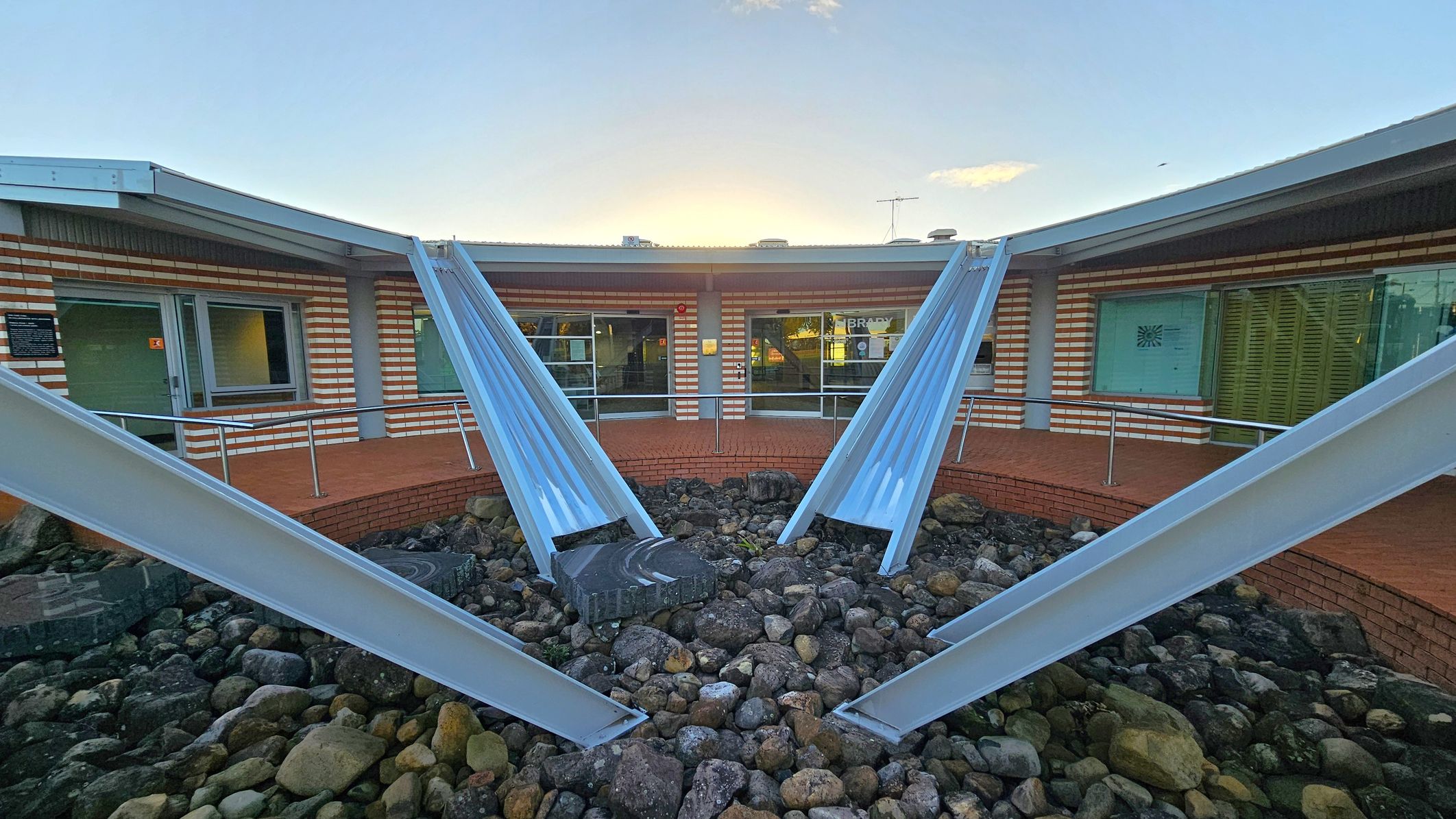 A courtyard with a circular arrangement of metal beams converging towards the centre, surrounded by a bed of rocks. The background features a building with a brick facade and large windows. The building has a flat roof with a metal edge. The ground is covered with stones, and there is a small raised brick platform near the building. The sky is visible above the structure. A courtyard with a circular arrangement of metal beams converging towards the centre, surrounded by a bed of rocks. The background features a building with a brick facade and large windows. The building has a flat roof with a metal edge. The ground is covered with stones, and there is a small raised brick platform near the building. The sky is visible above the structure.