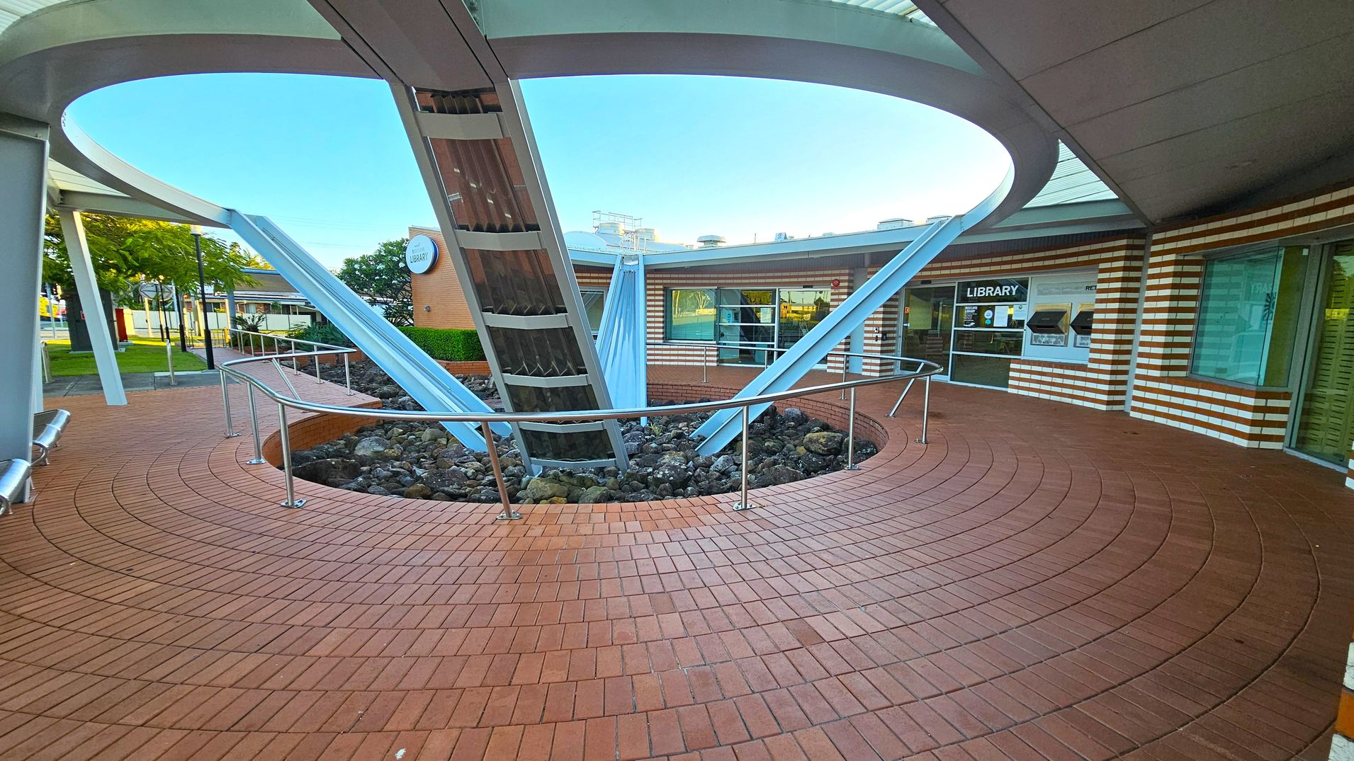 Circular courtyard with a brick floor and a central metal structure featuring angled beams. A railing surrounds a rocky area at the base of the structure. The background shows a building with striped brickwork and large windows, one marked "Library". Trees and a street are visible to the left. The ceiling is a curved metal canopy with an open circular section above the structure. Circular courtyard with a brick floor and a central metal structure featuring angled beams. A railing surrounds a rocky area at the base of the structure. The background shows a building with striped brickwork and large windows, one marked "Library". Trees and a street are visible to the left. The ceiling is a curved metal canopy with an open circular section above the structure.