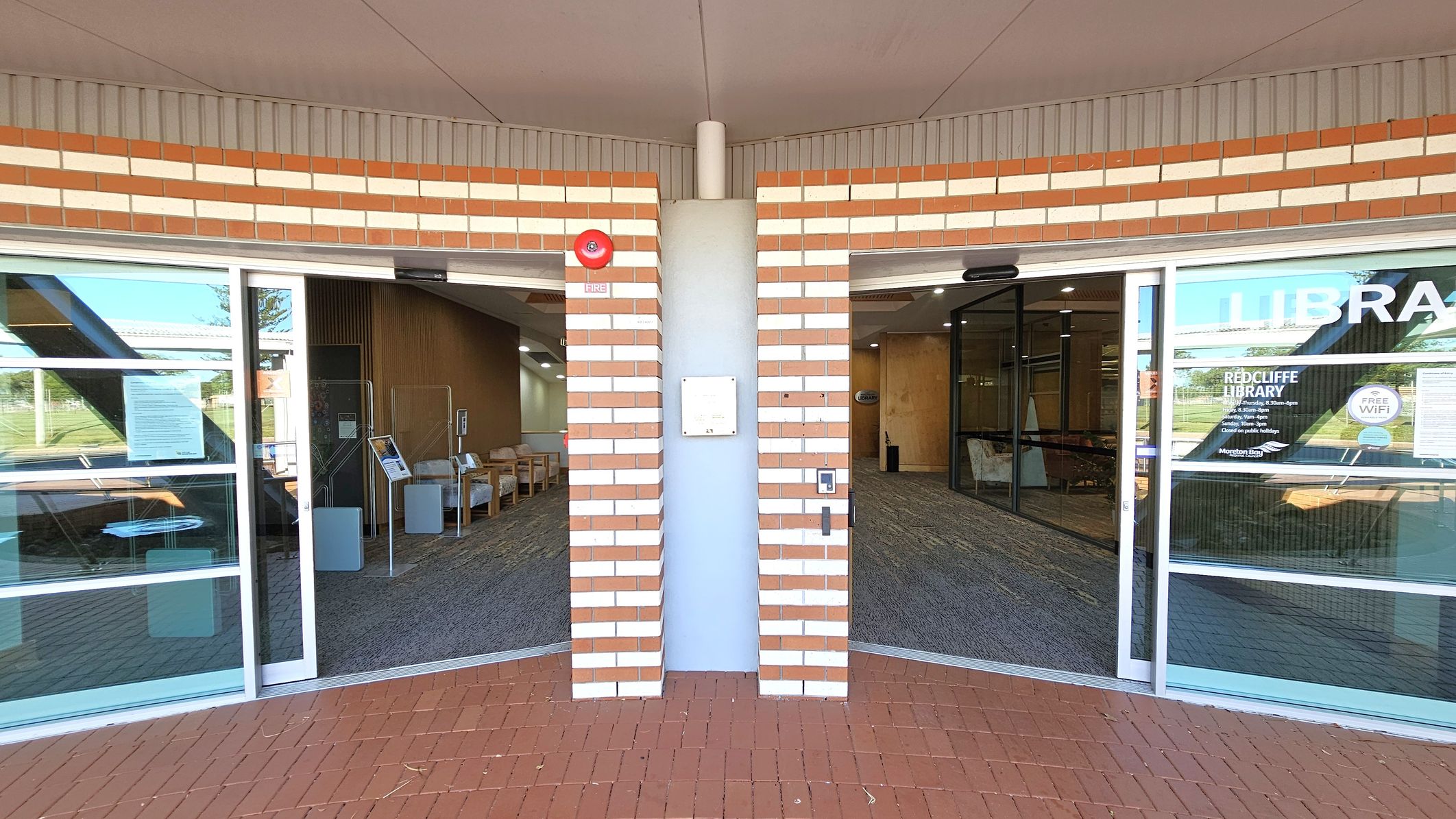 A brick wall with alternating red and white bricks is in the centre, featuring a fire alarm and a small sign. On either side, there are glass doors leading into a library. The left side shows a seating area with chairs and a notice board. The right side has signage for "Redcliffe Library" and "Free WiFi". The floor is covered with red tiles outside and a patterned carpet inside. The ceiling is plain with a central seam. A brick wall with alternating red and white bricks is in the centre, featuring a fire alarm and a small sign. On either side, there are glass doors leading into a library. The left side shows a seating area with chairs and a notice board. The right side has signage for "Redcliffe Library" and "Free WiFi". The floor is covered with red tiles outside and a patterned carpet inside. The ceiling is plain with a central seam.