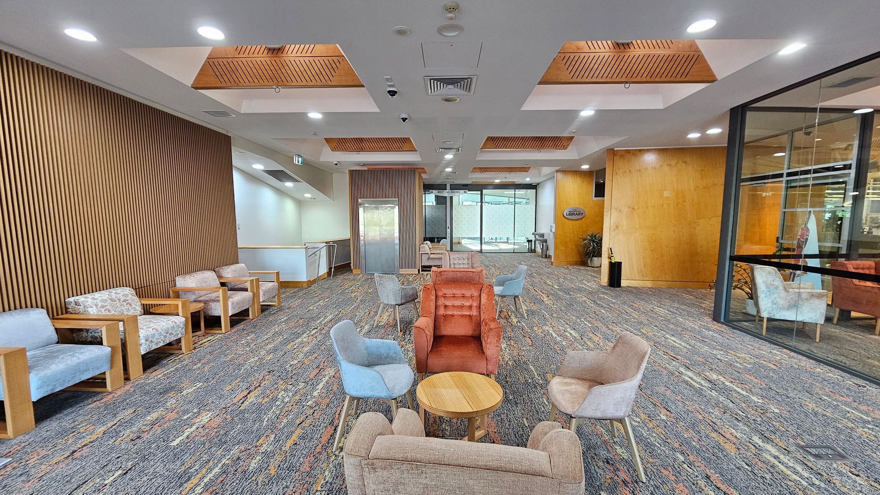A spacious lobby area with a patterned carpet floor. On the left, there is a row of cushioned chairs with wooden frames against a wall with vertical wooden slats. In the centre, a cluster of colourful armchairs surrounds small round tables. The ceiling features recessed lighting and wooden panels. On the right, a glass wall reveals more seating and a sign indicating a library. An elevator is visible in the background, flanked by more seating. A spacious lobby area with a patterned carpet floor. On the left, there is a row of cushioned chairs with wooden frames against a wall with vertical wooden slats. In the centre, a cluster of colourful armchairs surrounds small round tables. The ceiling features recessed lighting and wooden panels. On the right, a glass wall reveals more seating and a sign indicating a library. An elevator is visible in the background, flanked by more seating.