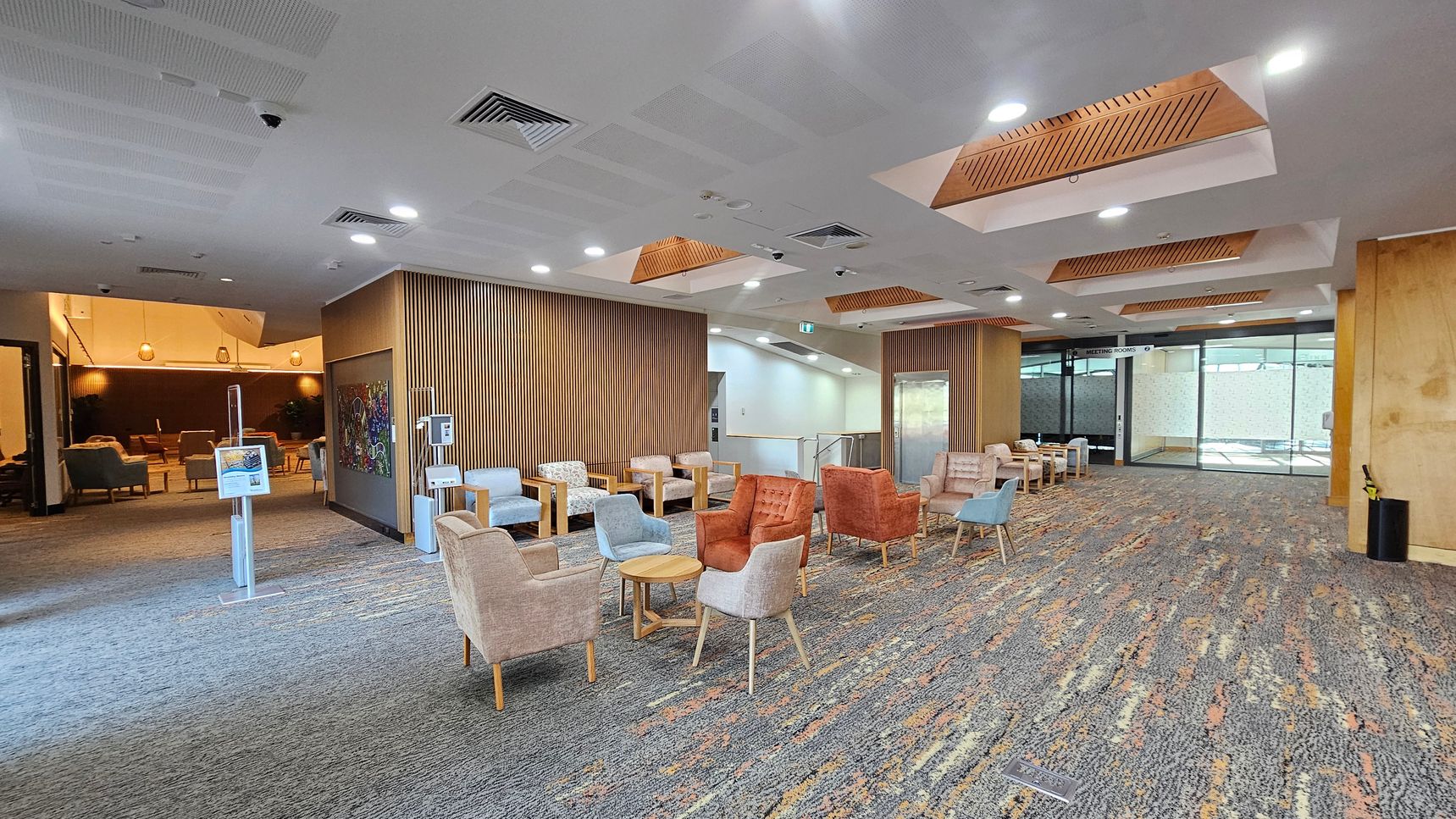 A spacious lobby area with a patterned carpet floor. On the left, there is a seating area with various chairs and small tables. The middle features a wooden panelled wall with a colourful artwork. On the right, more seating is arranged, and a glass door labelled "Meeting Rooms" is visible. The ceiling has recessed lighting and wooden accents. The background shows another seating area with warm lighting and pendant lamps. A spacious lobby area with a patterned carpet floor. On the left, there is a seating area with various chairs and small tables. The middle features a wooden panelled wall with a colourful artwork. On the right, more seating is arranged, and a glass door labelled "Meeting Rooms" is visible. The ceiling has recessed lighting and wooden accents. The background shows another seating area with warm lighting and pendant lamps.