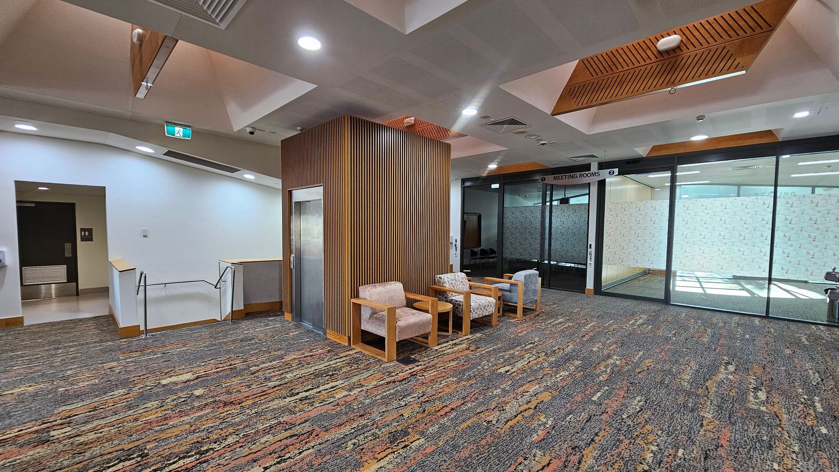A modern interior space with a patterned carpet floor. On the left, there's a staircase with a metal railing and a door with a sign above it. In the centre, a wooden panelled column houses a lift, with three cushioned chairs in front. On the right, a glass wall with a "Meeting Rooms" sign above it, partially frosted for privacy. The ceiling features recessed lighting and wooden accents. A modern interior space with a patterned carpet floor. On the left, there's a staircase with a metal railing and a door with a sign above it. In the centre, a wooden panelled column houses a lift, with three cushioned chairs in front. On the right, a glass wall with a "Meeting Rooms" sign above it, partially frosted for privacy. The ceiling features recessed lighting and wooden accents.