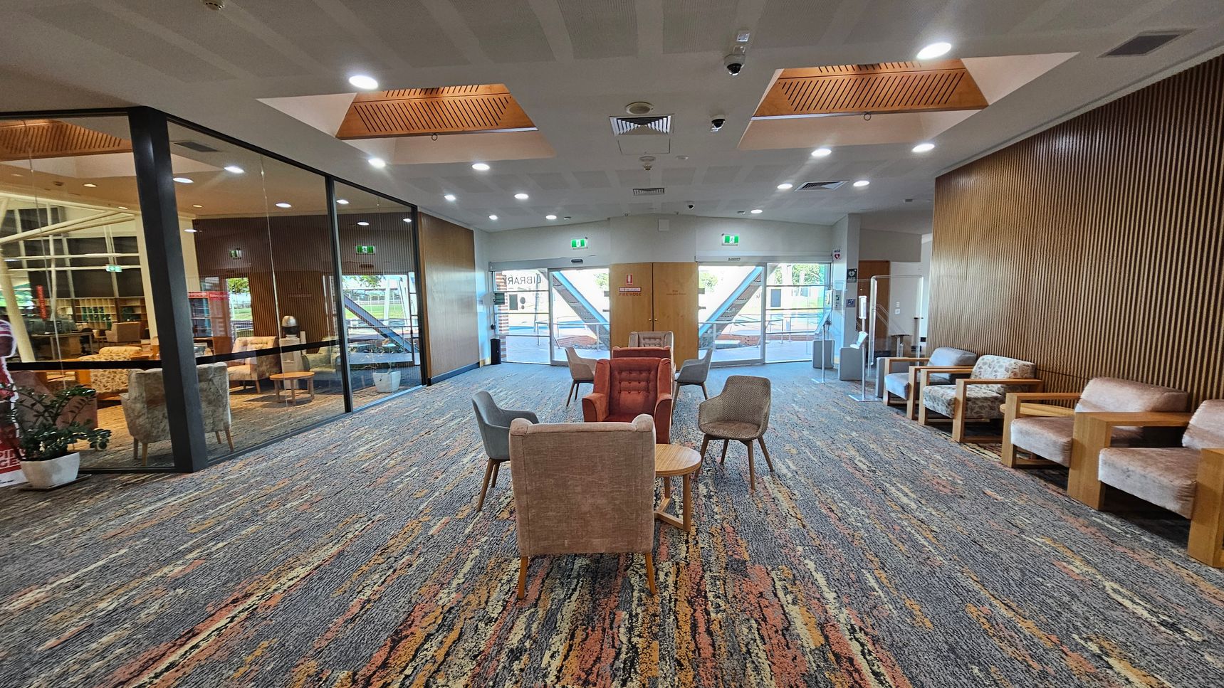 A spacious room with a patterned carpet in shades of grey, orange, and brown. In the centre, there are several chairs and small tables arranged in a casual seating area. The left side features a glass wall with a view into another room, while the right side has a wooden panelled wall with additional seating. The ceiling has recessed lighting and wooden accents. At the far end, there are large glass doors leading outside, flanked by emergency exit signs. A spacious room with a patterned carpet in shades of grey, orange, and brown. In the centre, there are several chairs and small tables arranged in a casual seating area. The left side features a glass wall with a view into another room, while the right side has a wooden panelled wall with additional seating. The ceiling has recessed lighting and wooden accents. At the far end, there are large glass doors leading outside, flanked by emergency exit signs.