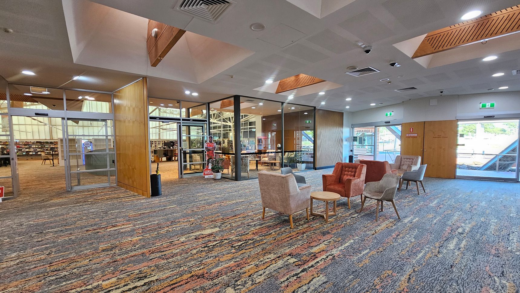 A spacious interior with a patterned carpet floor. On the right, there are several upholstered chairs and small round tables arranged in a seating area. The left side features glass doors leading to a library with visible bookshelves. The ceiling has recessed lighting and wooden accents. A cardboard cutout and potted plants are near the entrance. Large windows and doors provide natural light from outside. A spacious interior with a patterned carpet floor. On the right, there are several upholstered chairs and small round tables arranged in a seating area. The left side features glass doors leading to a library with visible bookshelves. The ceiling has recessed lighting and wooden accents. A cardboard cutout and potted plants are near the entrance. Large windows and doors provide natural light from outside.