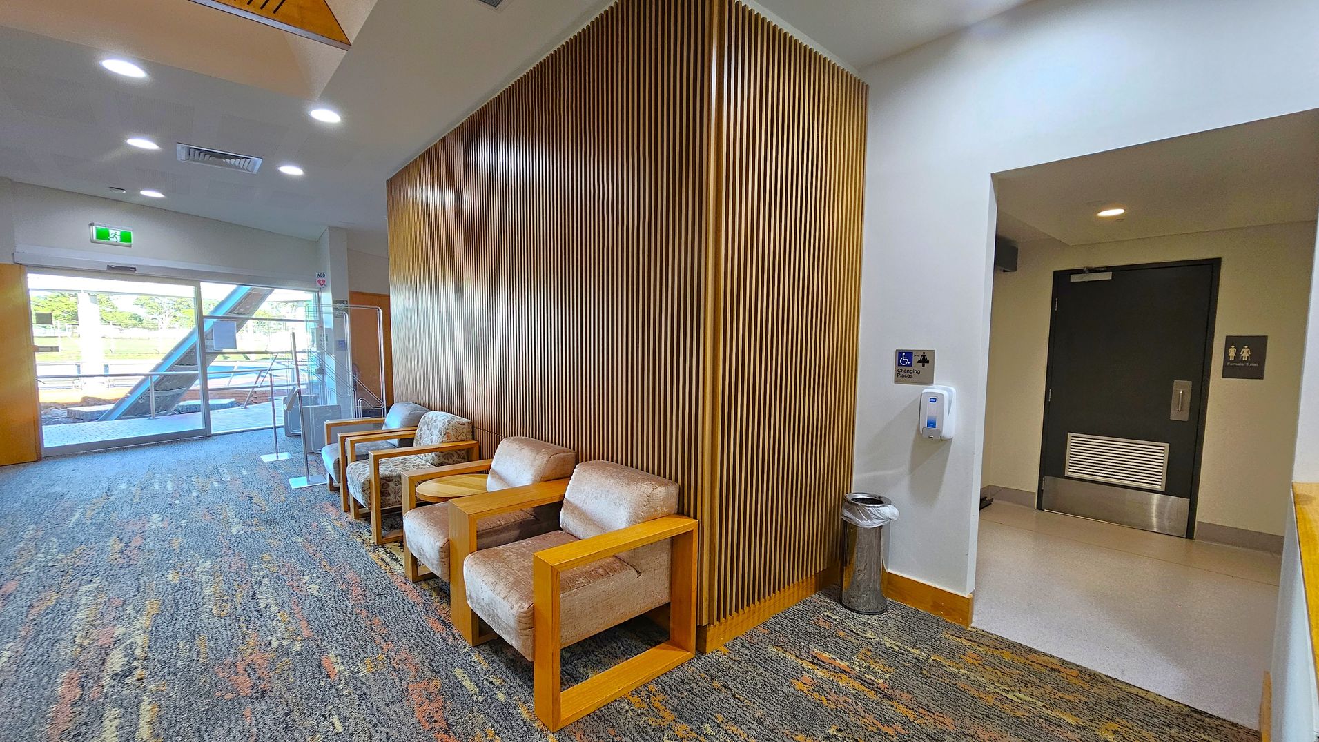 On the left, there is a glass door leading outside, with a green exit sign above. The floor is carpeted with a multicoloured pattern. In the middle, there are several cushioned chairs with wooden armrests against a wall with vertical wooden slats. On the right, a hallway leads to a dark door with restroom signs. A hand sanitiser dispenser and a small bin are mounted on the wall. The ceiling is white with recessed lighting. On the left, there is a glass door leading outside, with a green exit sign above. The floor is carpeted with a multicoloured pattern. In the middle, there are several cushioned chairs with wooden armrests against a wall with vertical wooden slats. On the right, a hallway leads to a dark door with restroom signs. A hand sanitiser dispenser and a small bin are mounted on the wall. The ceiling is white with recessed lighting.