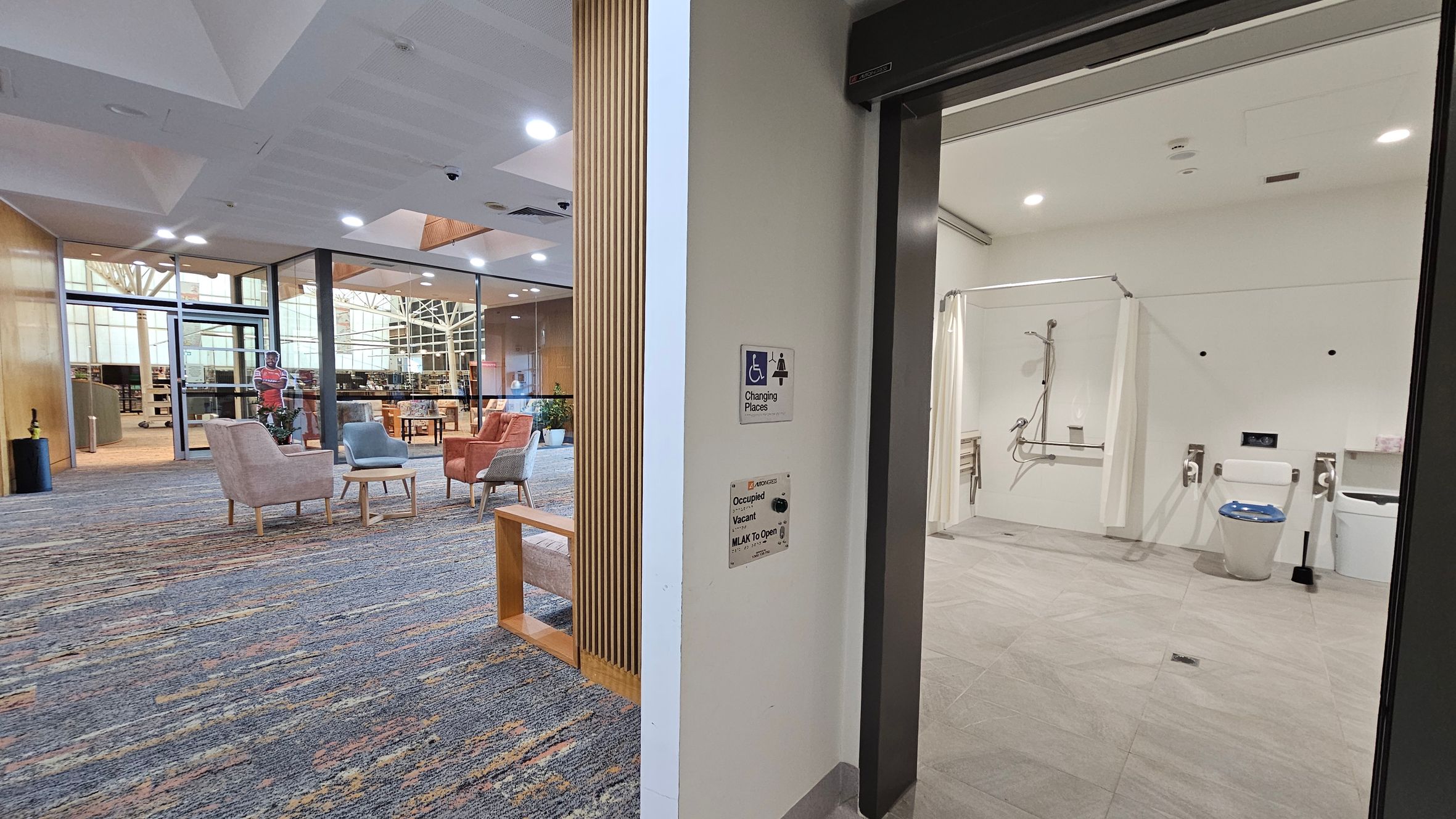 On the right, there's an accessible bathroom with a shower area, toilet, and support rails. The floor is tiled, and the ceiling has recessed lighting. On the left, a seating area with pastel-coloured chairs and small tables is visible, set on a patterned carpet. Beyond, glass walls reveal a library space with bookshelves and a cut-out figure. The ceiling is white with embedded lights, and a wooden partition partially separates the two areas. On the right, there's an accessible bathroom with a shower area, toilet, and support rails. The floor is tiled, and the ceiling has recessed lighting. On the left, a seating area with pastel-coloured chairs and small tables is visible, set on a patterned carpet. Beyond, glass walls reveal a library space with bookshelves and a cut-out figure. The ceiling is white with embedded lights, and a wooden partition partially separates the two areas.