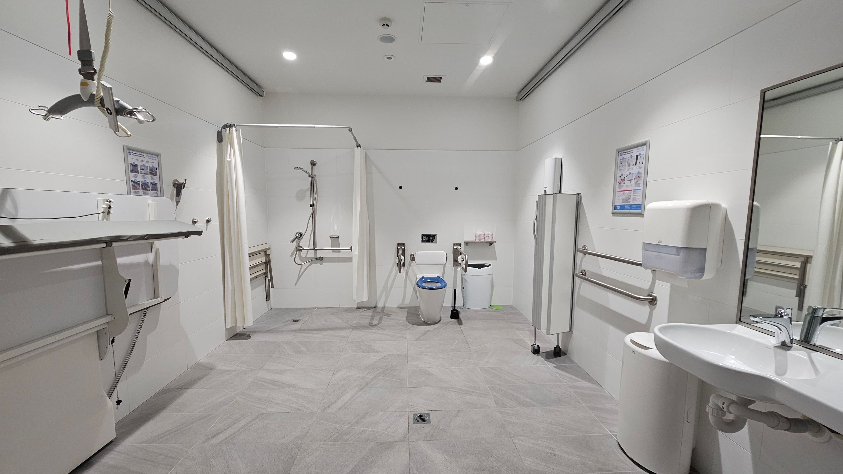A spacious, accessible bathroom with grey tiled flooring and white walls. On the left, there is a height-adjustable changing table and a ceiling hoist. In the centre, a shower area with a curtain and a toilet equipped with support rails. On the right, a sink with a mirror above, a soap dispenser, and a waste bin. The ceiling is fitted with recessed lighting. Various informational posters are mounted on the walls. A spacious, accessible bathroom with grey tiled flooring and white walls. On the left, there is a height-adjustable changing table and a ceiling hoist. In the centre, a shower area with a curtain and a toilet equipped with support rails. On the right, a sink with a mirror above, a soap dispenser, and a waste bin. The ceiling is fitted with recessed lighting. Various informational posters are mounted on the walls.
