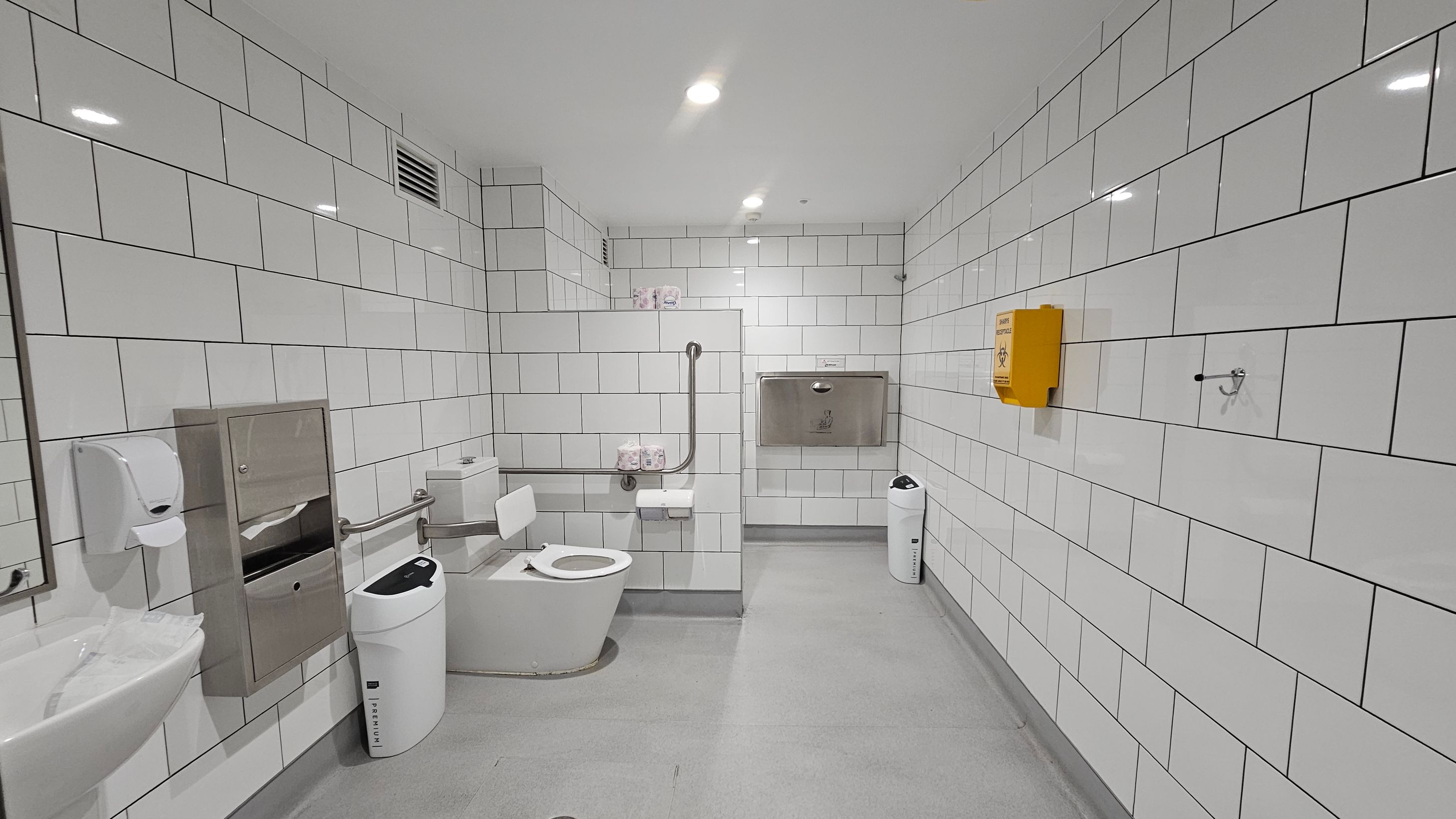 A clean, accessible bathroom with white tiled walls and a light grey floor. On the left, there's a sink with a soap dispenser and a paper towel holder. A toilet with support rails is positioned centrally, with toilet paper and a sanitary bin nearby. On the right, a yellow biohazard disposal box is mounted on the wall. The ceiling is white with recessed lighting, and a baby changing station is visible on the back wall. A clean, accessible bathroom with white tiled walls and a light grey floor. On the left, there's a sink with a soap dispenser and a paper towel holder. A toilet with support rails is positioned centrally, with toilet paper and a sanitary bin nearby. On the right, a yellow biohazard disposal box is mounted on the wall. The ceiling is white with recessed lighting, and a baby changing station is visible on the back wall.