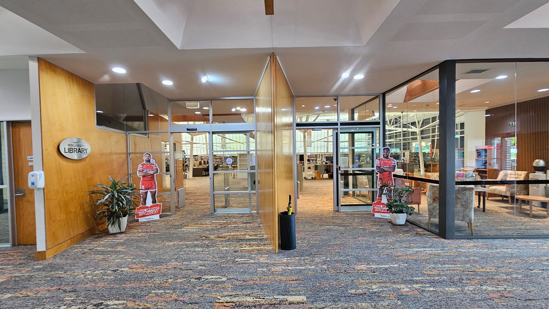 Entrance to a library with a glass door in the centre, flanked by wooden panels. On the left, a sign reads "Library" above a sanitiser dispenser and a potted plant. On the right, a similar potted plant and a cardboard cut-out of a person in a red outfit. Inside, bookshelves and seating areas are visible. The floor is carpeted with a multicoloured pattern, and the ceiling has recessed lighting. Entrance to a library with a glass door in the centre, flanked by wooden panels. On the left, a sign reads "Library" above a sanitiser dispenser and a potted plant. On the right, a similar potted plant and a cardboard cut-out of a person in a red outfit. Inside, bookshelves and seating areas are visible. The floor is carpeted with a multicoloured pattern, and the ceiling has recessed lighting.