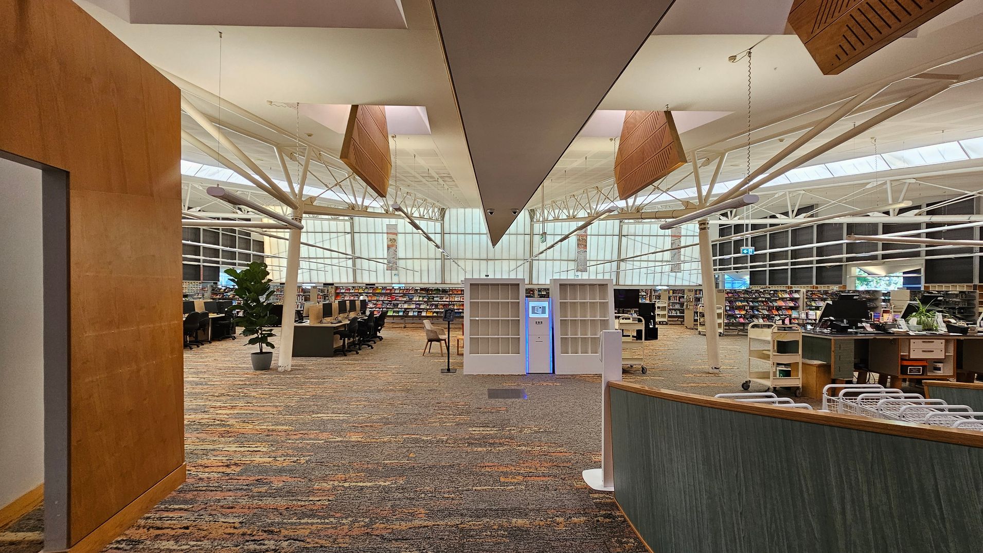 A spacious library interior with a high ceiling featuring exposed beams and large windows in the background. In the centre, there are two white shelving units with a digital kiosk between them. To the left, there are rows of computer stations and a potted plant. On the right, a curved desk with baskets and office supplies. The floor is covered with a patterned carpet in earthy tones. The ceiling has suspended wooden panels and modern lighting fixtures. A spacious library interior with a high ceiling featuring exposed beams and large windows in the background. In the centre, there are two white shelving units with a digital kiosk between them. To the left, there are rows of computer stations and a potted plant. On the right, a curved desk with baskets and office supplies. The floor is covered with a patterned carpet in earthy tones. The ceiling has suspended wooden panels and modern lighting fixtures.