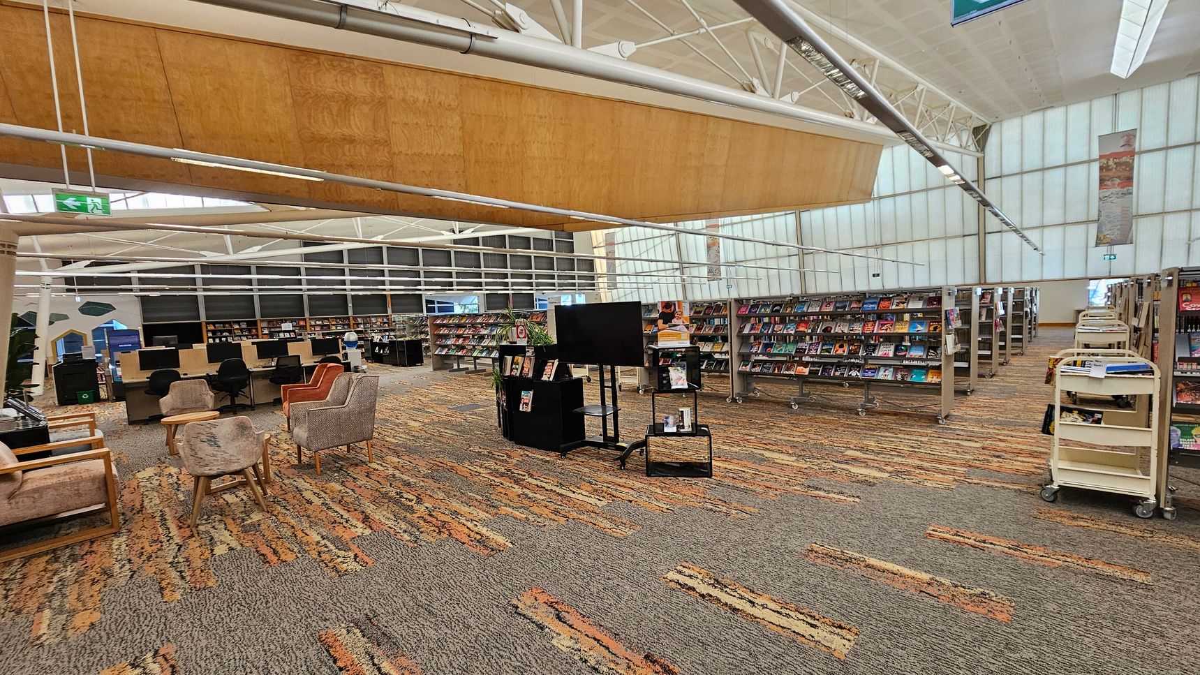 A spacious library interior with a patterned carpet floor. On the left, there are several comfortable chairs and computer stations with black chairs. In the centre, a black stand holds a large screen and some books. To the right, rows of bookshelves are filled with books, and there are book carts nearby. The ceiling is high with exposed beams and a large wooden panel. The walls are made of translucent panels, allowing natural light to filter through. A spacious library interior with a patterned carpet floor. On the left, there are several comfortable chairs and computer stations with black chairs. In the centre, a black stand holds a large screen and some books. To the right, rows of bookshelves are filled with books, and there are book carts nearby. The ceiling is high with exposed beams and a large wooden panel. The walls are made of translucent panels, allowing natural light to filter through.