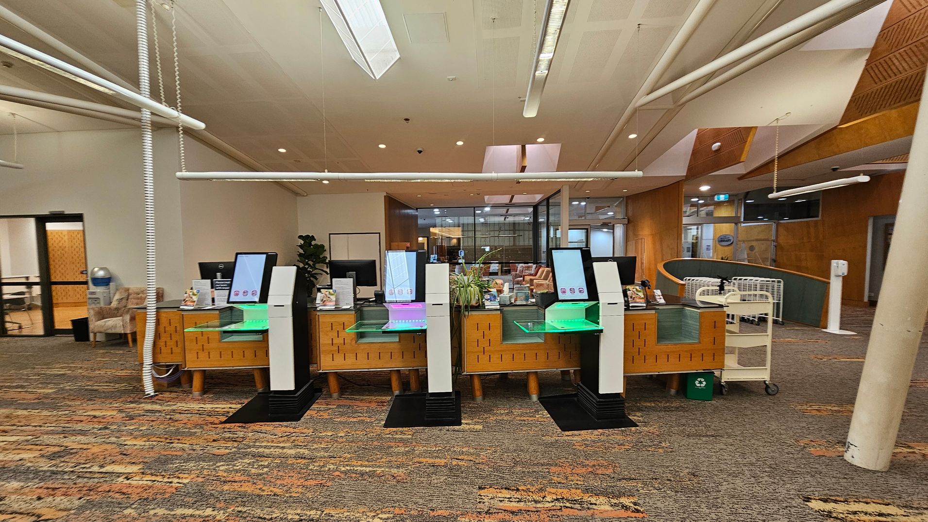 A library interior with a row of self-service kiosks in the centre, each with a screen and a glowing base. Behind them is a wooden counter with various items and a plant. To the left, there's a seating area with a water cooler and chairs. On the right, a book cart and a recycling bin are visible. The floor is carpeted with a multicoloured pattern, and the ceiling features white panels and lights. The background includes glass doors and wooden walls. A library interior with a row of self-service kiosks in the centre, each with a screen and a glowing base. Behind them is a wooden counter with various items and a plant. To the left, there's a seating area with a water cooler and chairs. On the right, a book cart and a recycling bin are visible. The floor is carpeted with a multicoloured pattern, and the ceiling features white panels and lights. The background includes glass doors and wooden walls.