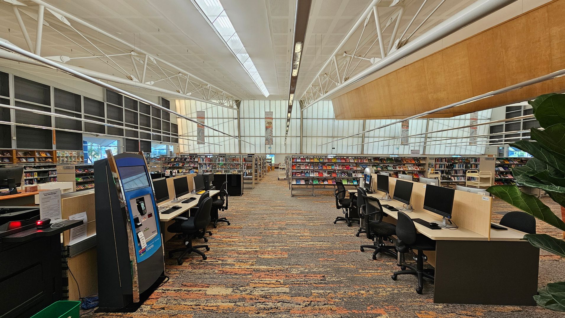 A spacious library interior with a high ceiling featuring exposed white beams. In the foreground, rows of computer stations with black chairs are on both sides. The floor is covered with a patterned carpet in earthy tones. In the background, bookshelves filled with colourful books line the walls. Large windows on the left allow natural light to illuminate the space. A large plant is partially visible on the right. A spacious library interior with a high ceiling featuring exposed white beams. In the foreground, rows of computer stations with black chairs are on both sides. The floor is covered with a patterned carpet in earthy tones. In the background, bookshelves filled with colourful books line the walls. Large windows on the left allow natural light to illuminate the space. A large plant is partially visible on the right.
