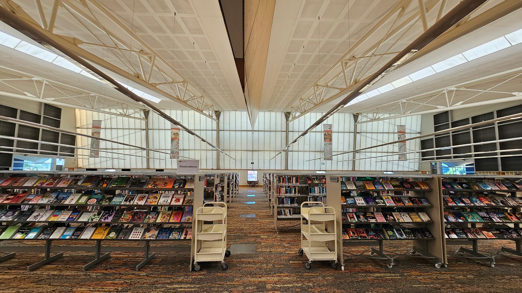 Bookshelves filled with various books are in the foreground, with two white book carts positioned in front. The background features large, translucent windows allowing natural light to illuminate the space. The ceiling is high with visible metal beams and a grid pattern. The floor is carpeted with a multicoloured pattern. The overall layout is spacious and organised, with a clear path between the shelves. Bookshelves filled with various books are in the foreground, with two white book carts positioned in front. The background features large, translucent windows allowing natural light to illuminate the space. The ceiling is high with visible metal beams and a grid pattern. The floor is carpeted with a multicoloured pattern. The overall layout is spacious and organised, with a clear path between the shelves.