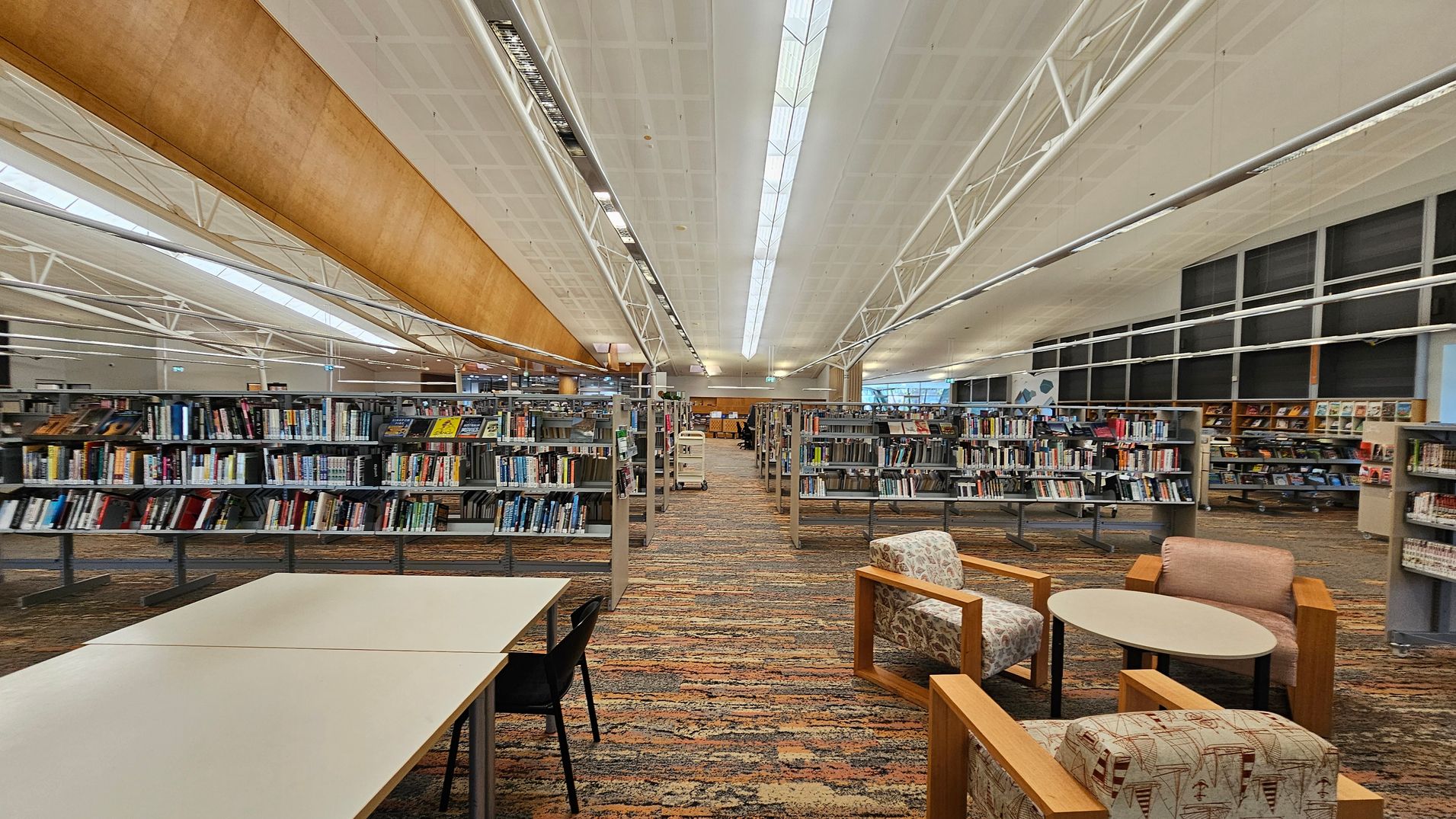 A spacious library interior with rows of bookshelves filled with books in the centre. On the left, there are white tables with black chairs. On the right, there are cushioned chairs with patterned upholstery and a small round table. The floor is covered with a multicoloured carpet. The ceiling is high with white panels and long fluorescent lights. Large windows are visible in the background, allowing natural light to enter. A spacious library interior with rows of bookshelves filled with books in the centre. On the left, there are white tables with black chairs. On the right, there are cushioned chairs with patterned upholstery and a small round table. The floor is covered with a multicoloured carpet. The ceiling is high with white panels and long fluorescent lights. Large windows are visible in the background, allowing natural light to enter.