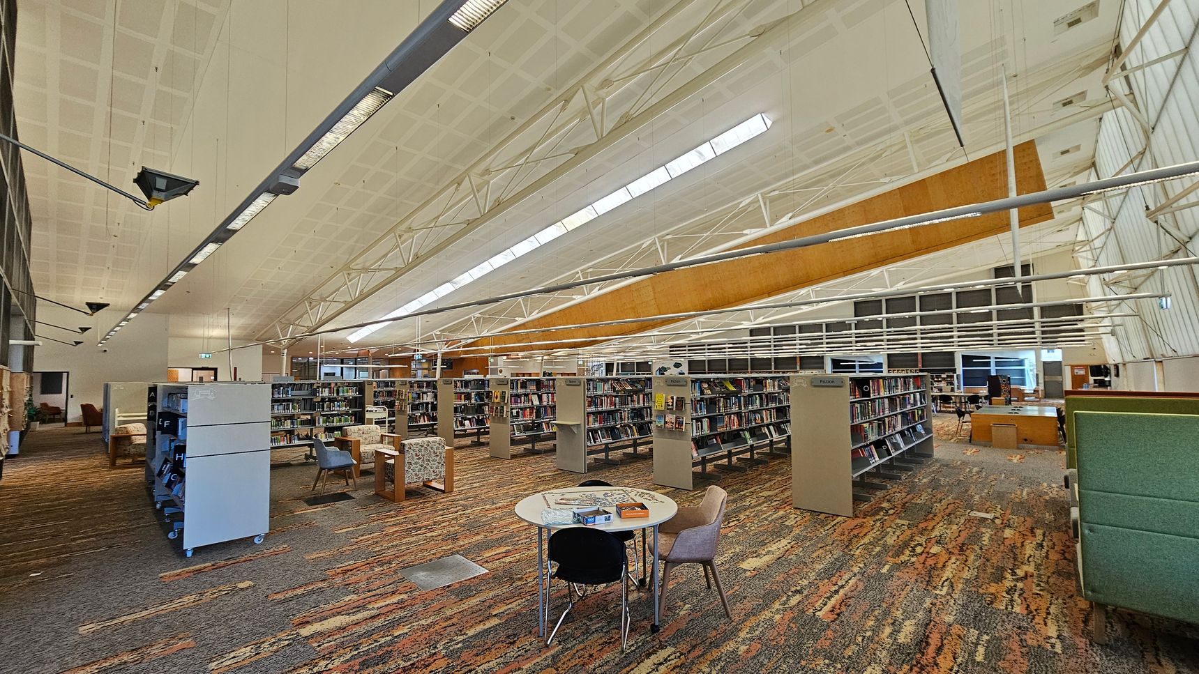 A spacious library interior with a high, white ceiling featuring exposed beams and skylights. In the foreground, a round table with chairs is placed on a multicoloured carpet. Bookshelves filled with books are arranged in rows across the room. On the left, there are more bookshelves and a few armchairs. The right side has additional seating and a green partition. The background shows large windows and more tables and chairs. The lighting is bright and evenly distributed. A spacious library interior with a high, white ceiling featuring exposed beams and skylights. In the foreground, a round table with chairs is placed on a multicoloured carpet. Bookshelves filled with books are arranged in rows across the room. On the left, there are more bookshelves and a few armchairs. The right side has additional seating and a green partition. The background shows large windows and more tables and chairs. The lighting is bright and evenly distributed.