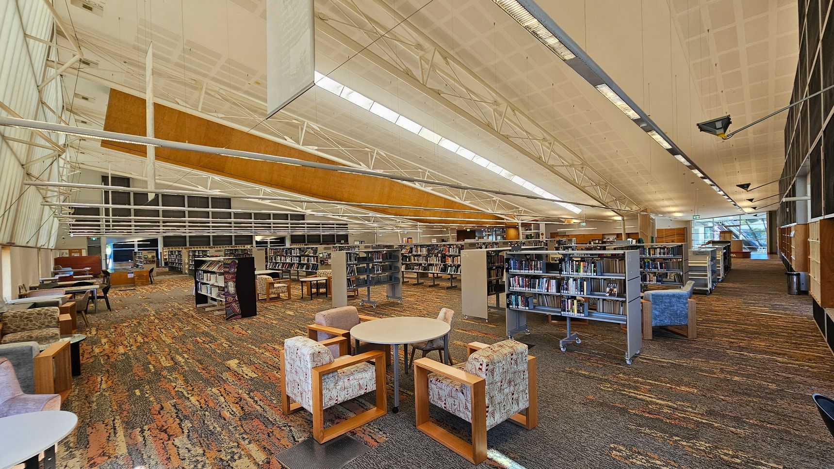 A spacious library interior with a high, white ceiling featuring exposed beams and skylights. In the foreground, there are round tables surrounded by patterned armchairs. The floor is covered with a multicoloured carpet. Bookshelves filled with books are arranged in rows across the middle and background. On the left, there are additional seating areas with tables and chairs. The right side has more bookshelves and a large window allowing natural light to enter. A spacious library interior with a high, white ceiling featuring exposed beams and skylights. In the foreground, there are round tables surrounded by patterned armchairs. The floor is covered with a multicoloured carpet. Bookshelves filled with books are arranged in rows across the middle and background. On the left, there are additional seating areas with tables and chairs. The right side has more bookshelves and a large window allowing natural light to enter.