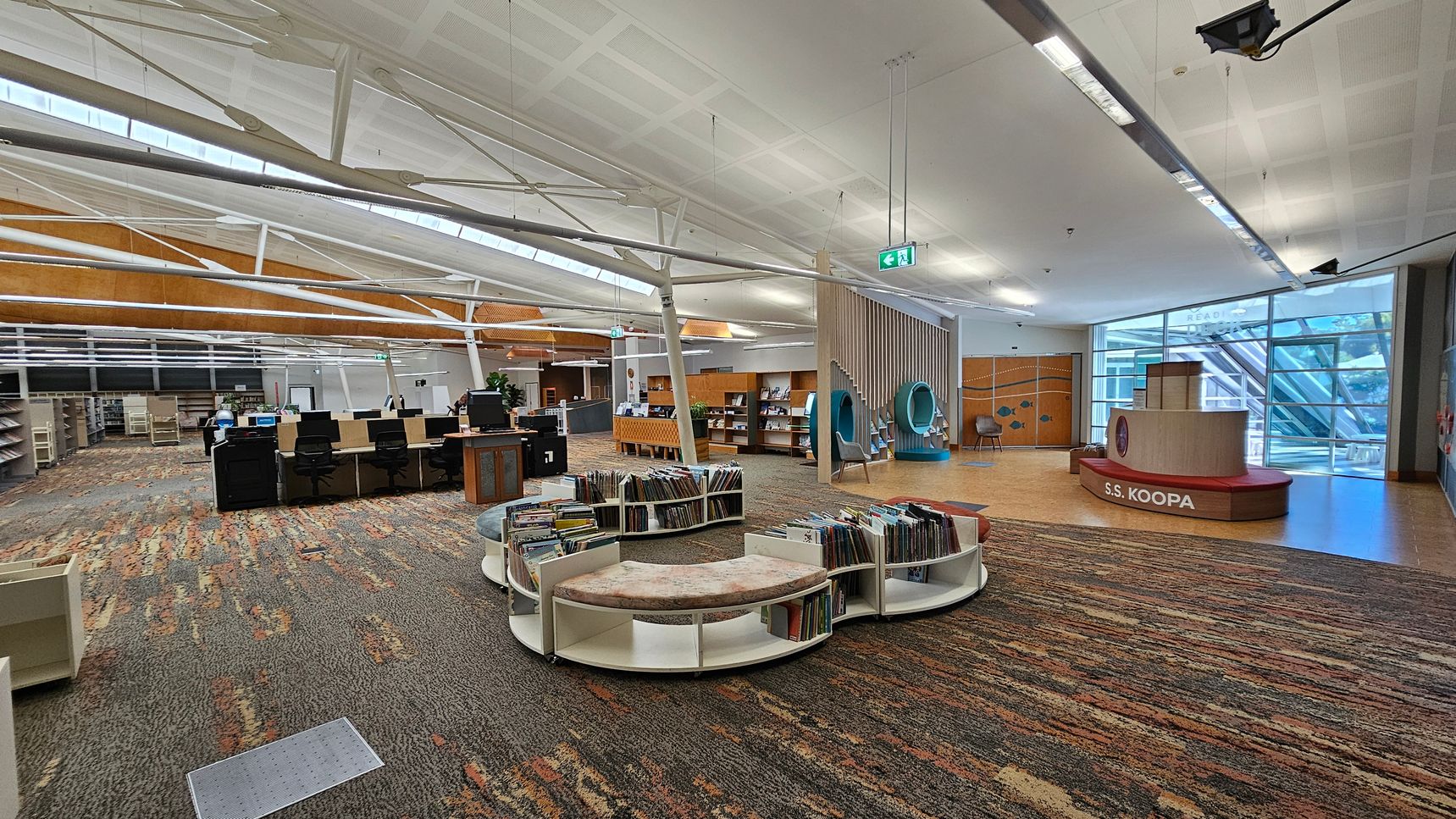 A spacious library interior with a colourful, patterned carpet. On the left, there are rows of bookshelves and a line of computer desks with chairs. In the centre, circular bookshelves with cushioned seating display books. On the right, a play area features a structure labelled "S.S. KOOPA" and two circular reading nooks. The ceiling is high with exposed beams, and large windows in the background allow natural light to enter. A spacious library interior with a colourful, patterned carpet. On the left, there are rows of bookshelves and a line of computer desks with chairs. In the centre, circular bookshelves with cushioned seating display books. On the right, a play area features a structure labelled "S.S. KOOPA" and two circular reading nooks. The ceiling is high with exposed beams, and large windows in the background allow natural light to enter.