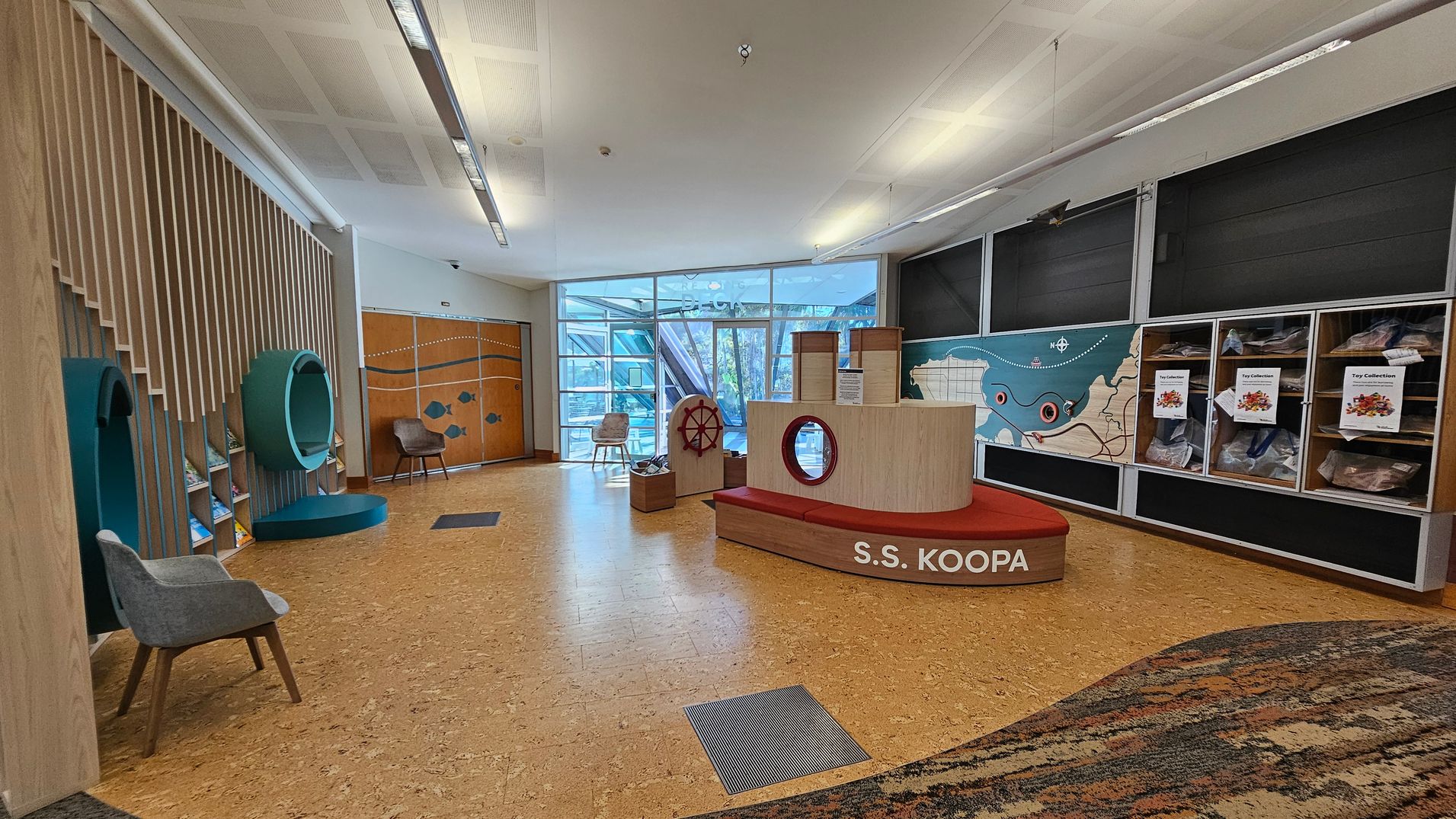 A spacious room with a cork floor and a white ceiling. On the left, there are wooden slats with circular green structures and a grey chair. In the centre, a ship-themed play area labelled "S.S. KOOPA" with a red bench and a ship's wheel. The right side features a wall with a map and display cases labelled "Toy Collection." The background has large windows allowing natural light, and a wooden door with fish designs. A spacious room with a cork floor and a white ceiling. On the left, there are wooden slats with circular green structures and a grey chair. In the centre, a ship-themed play area labelled "S.S. KOOPA" with a red bench and a ship's wheel. The right side features a wall with a map and display cases labelled "Toy Collection." The background has large windows allowing natural light, and a wooden door with fish designs.
