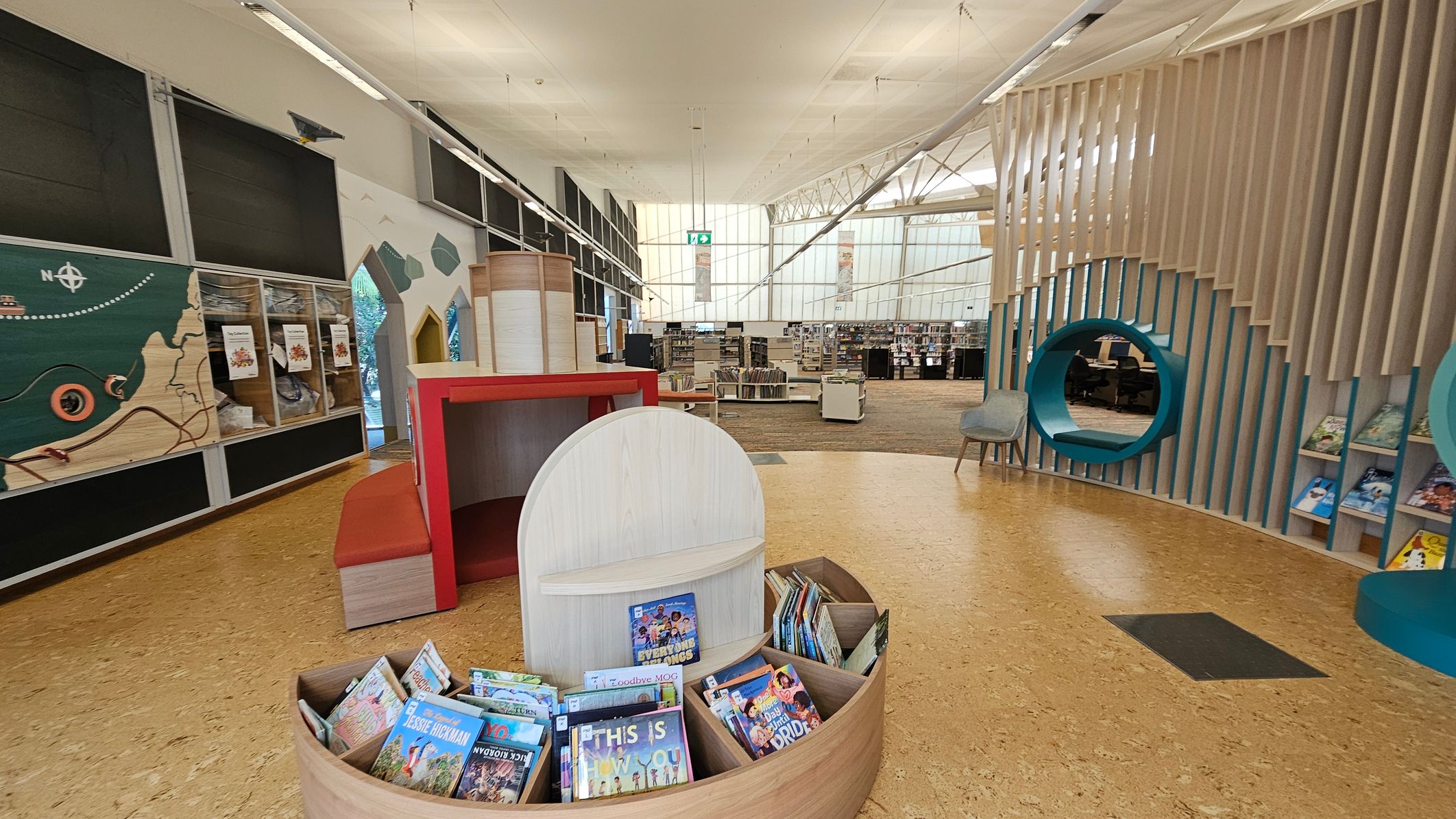 A modern library interior with a cork floor. On the left, a wall features a map design and display cases. In the centre, a circular wooden book display holds children's books. The background shows bookshelves and a large window with a grid pattern. On the right, a seating area with a circular cut-out in a slatted wall, accompanied by a chair. The ceiling is high with exposed beams and lighting fixtures. A modern library interior with a cork floor. On the left, a wall features a map design and display cases. In the centre, a circular wooden book display holds children's books. The background shows bookshelves and a large window with a grid pattern. On the right, a seating area with a circular cut-out in a slatted wall, accompanied by a chair. The ceiling is high with exposed beams and lighting fixtures.