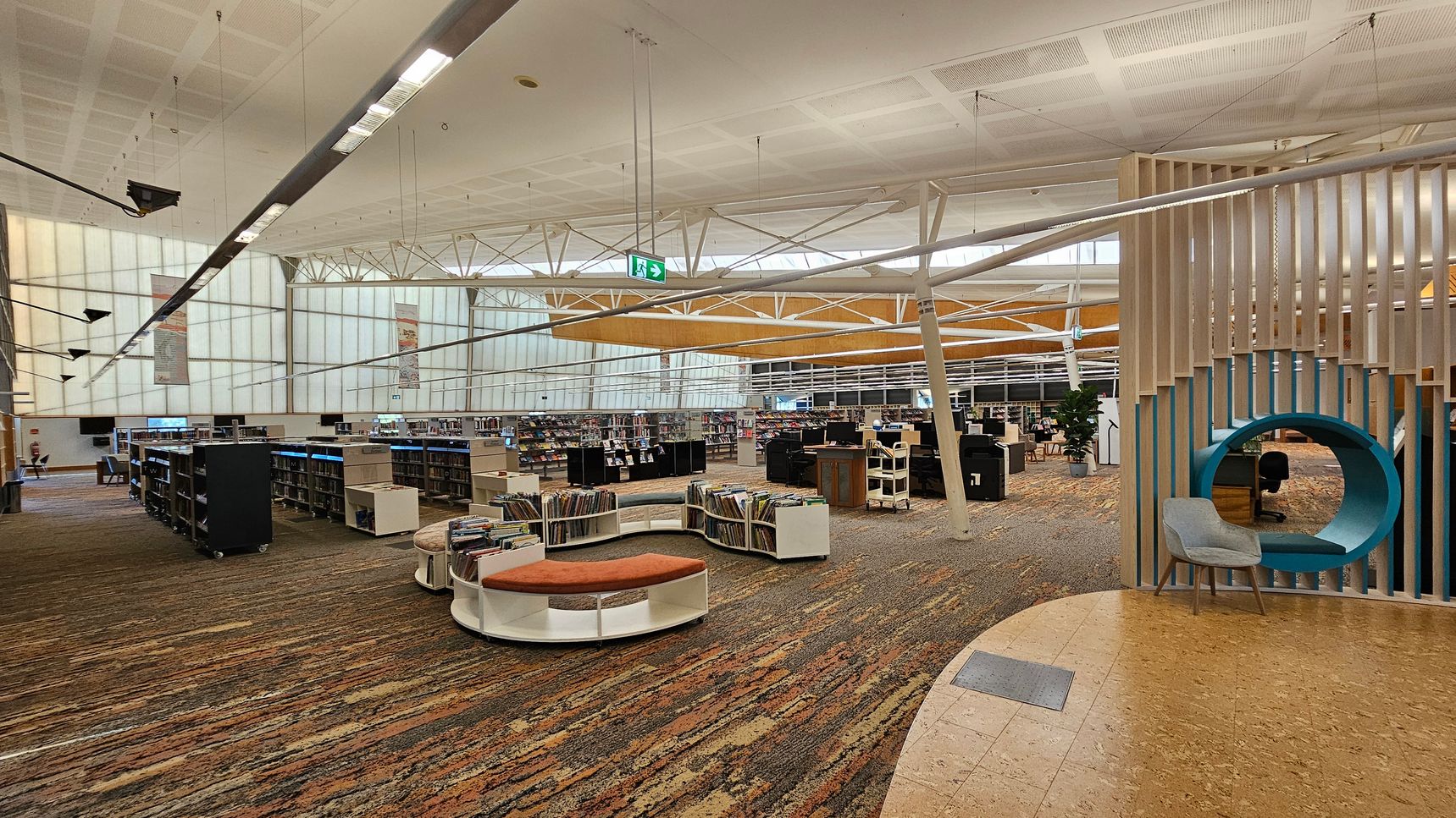 A spacious library interior with a high, white ceiling featuring exposed beams and hanging lights. On the left, there are rows of bookshelves filled with books. In the centre, a circular seating area with orange cushions is surrounded by more bookshelves. On the right, a unique seating nook with a blue circular frame and a grey chair is integrated into a wooden slat partition. The floor is covered with a patterned carpet, and a section of cork flooring is visible in the foreground. A spacious library interior with a high, white ceiling featuring exposed beams and hanging lights. On the left, there are rows of bookshelves filled with books. In the centre, a circular seating area with orange cushions is surrounded by more bookshelves. On the right, a unique seating nook with a blue circular frame and a grey chair is integrated into a wooden slat partition. The floor is covered with a patterned carpet, and a section of cork flooring is visible in the foreground.