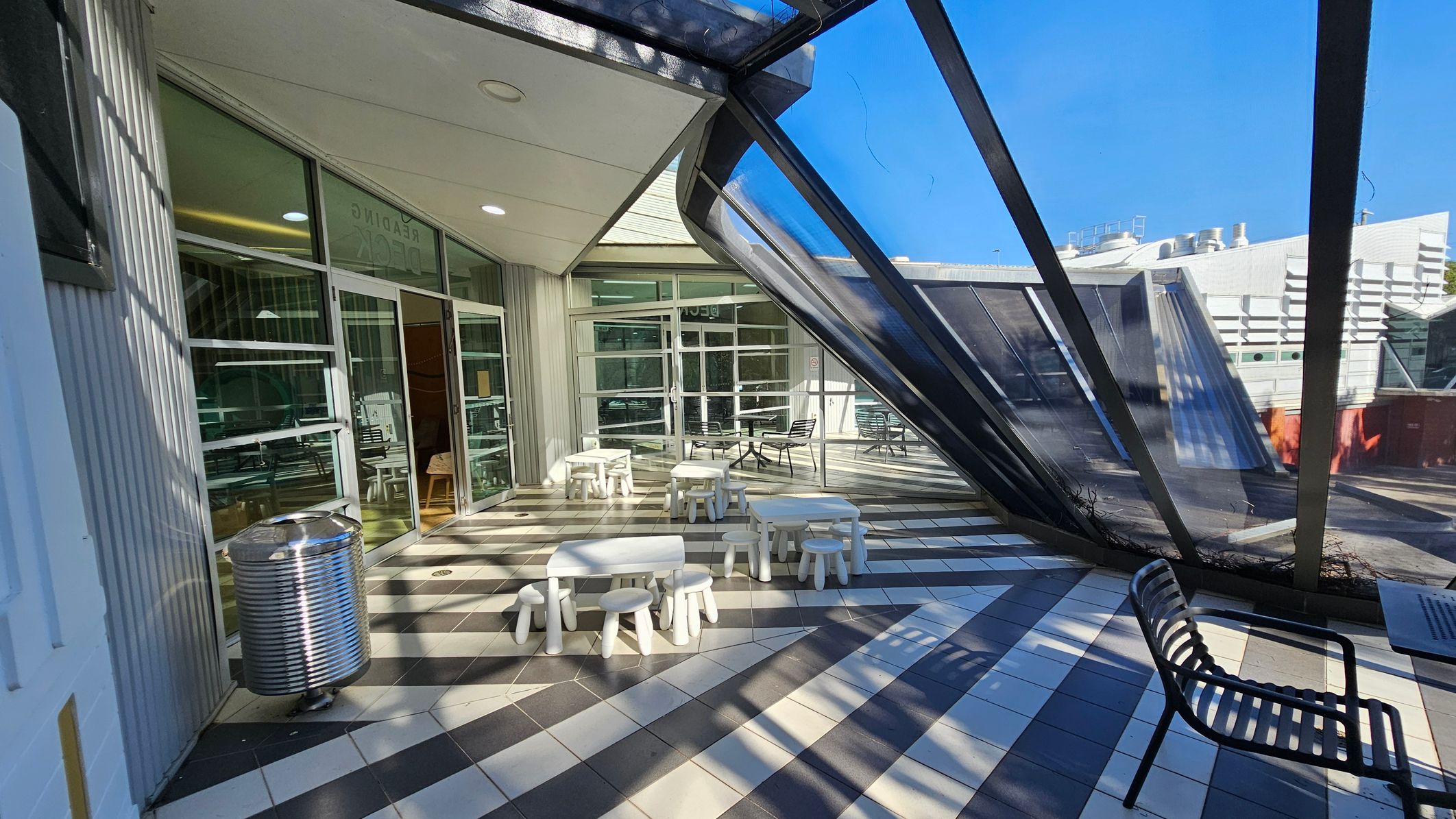 A modern indoor space with a slanted glass wall on the right, allowing natural light to flood in. The floor is tiled in a black and white checker pattern. In the centre, there are small white tables and stools arranged in a casual seating area. On the left, a metal bin is placed near glass doors leading to another room. A black metal bench is positioned on the right side. The ceiling is white with recessed lighting. A modern indoor space with a slanted glass wall on the right, allowing natural light to flood in. The floor is tiled in a black and white checker pattern. In the centre, there are small white tables and stools arranged in a casual seating area. On the left, a metal bin is placed near glass doors leading to another room. A black metal bench is positioned on the right side. The ceiling is white with recessed lighting.