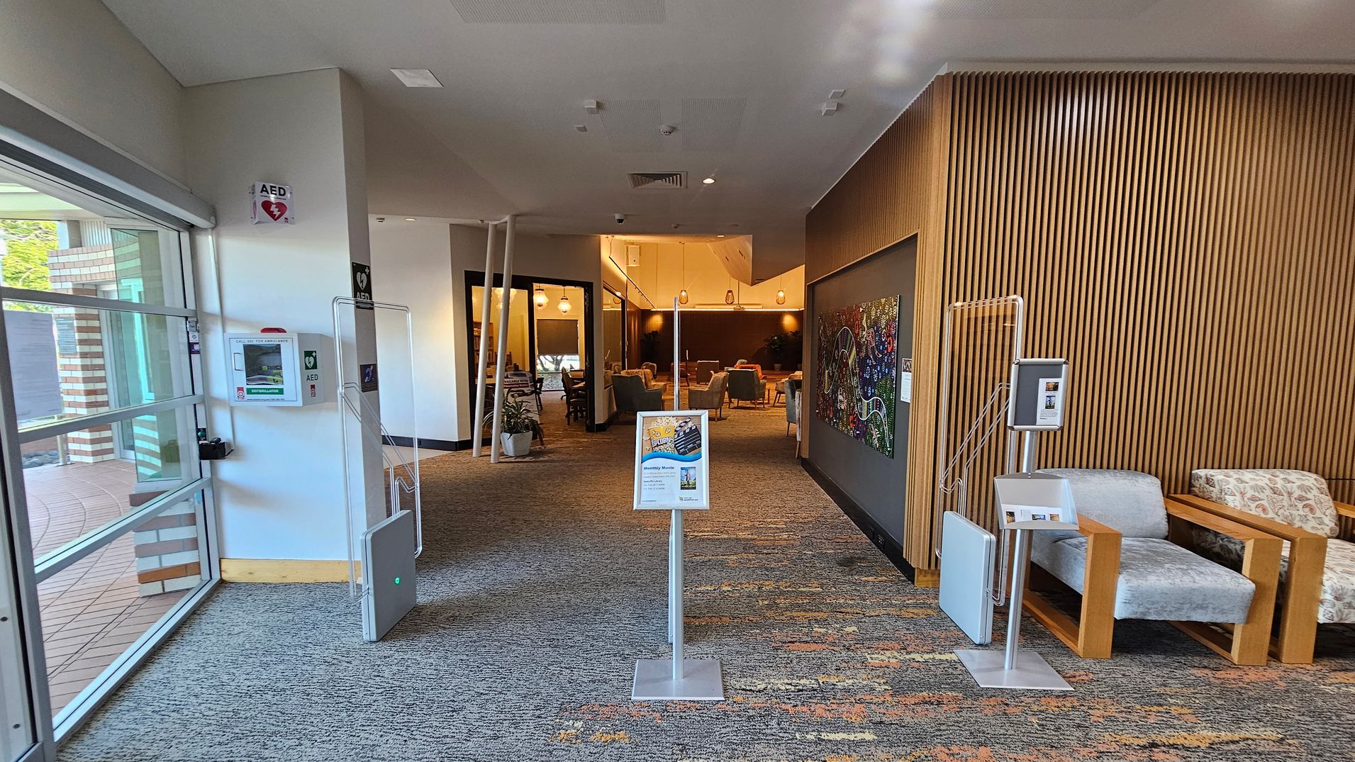 On the left, a glass wall with a view of the outside and an AED mounted on the wall. In the centre, a stand with a sign and a view into a seating area with tables and chairs. The right side features a wooden slatted wall with a colourful artwork and two cushioned chairs with wooden frames. The floor is carpeted with a multicoloured pattern, and the ceiling is white with recessed lighting. On the left, a glass wall with a view of the outside and an AED mounted on the wall. In the centre, a stand with a sign and a view into a seating area with tables and chairs. The right side features a wooden slatted wall with a colourful artwork and two cushioned chairs with wooden frames. The floor is carpeted with a multicoloured pattern, and the ceiling is white with recessed lighting.