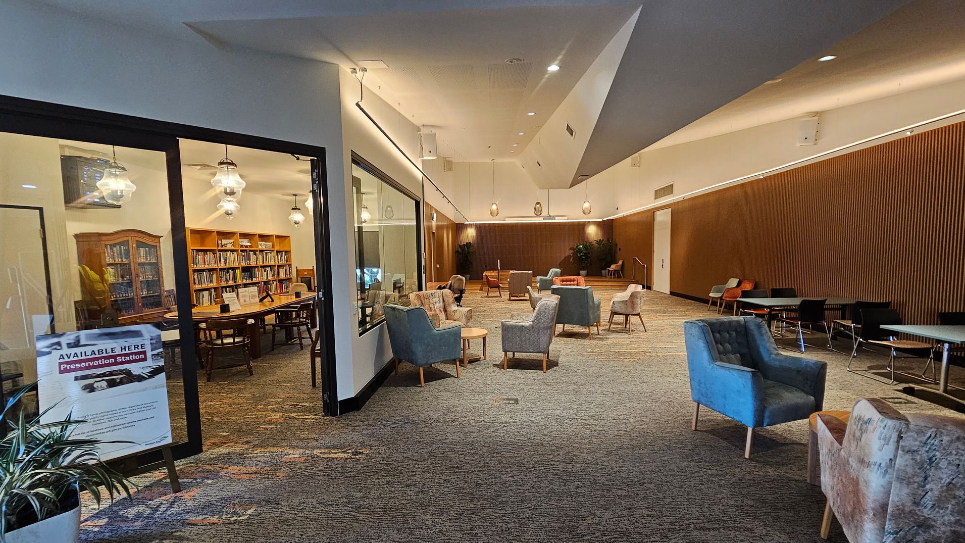 A spacious interior with a library room on the left, visible through a glass wall. The library has wooden bookshelves filled with books and a large table with chairs. A sign outside reads "Available Here Preservation Station." The main area has a variety of seating, including armchairs and small tables, on a patterned carpet. The ceiling is high with modern lighting, and the far wall is wooden with plants placed at intervals. A spacious interior with a library room on the left, visible through a glass wall. The library has wooden bookshelves filled with books and a large table with chairs. A sign outside reads "Available Here Preservation Station." The main area has a variety of seating, including armchairs and small tables, on a patterned carpet. The ceiling is high with modern lighting, and the far wall is wooden with plants placed at intervals.