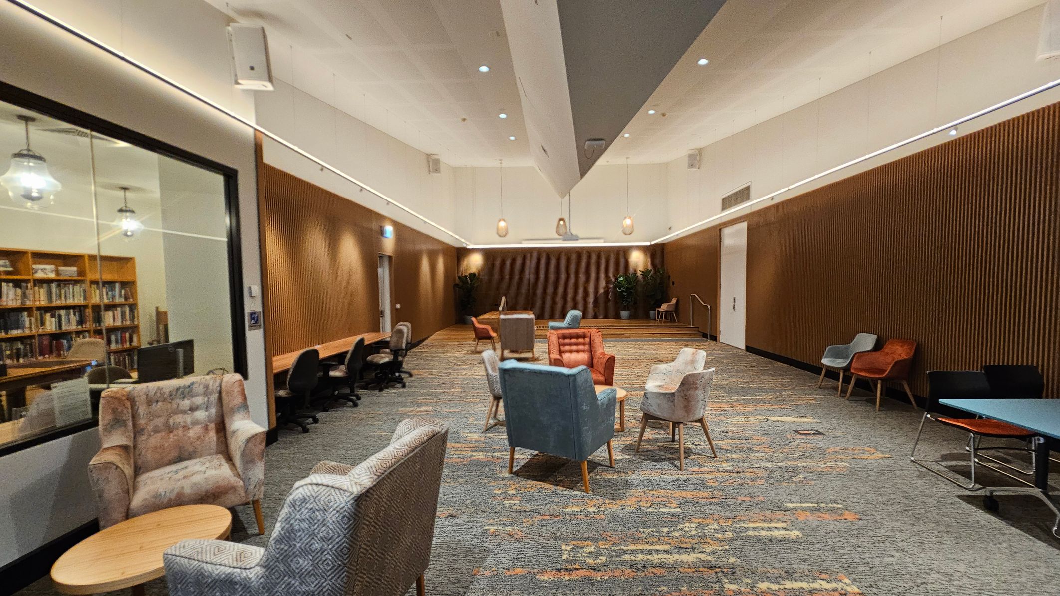 A spacious room with a high ceiling and modern lighting. On the left, there's a glass wall revealing a library with bookshelves. In the centre, a cluster of colourful armchairs surrounds small tables on a patterned carpet. The right side features a wooden panelled wall with additional seating, including chairs and a table. The floor is carpeted with a mix of green and orange tones, and there are potted plants in the background. A spacious room with a high ceiling and modern lighting. On the left, there's a glass wall revealing a library with bookshelves. In the centre, a cluster of colourful armchairs surrounds small tables on a patterned carpet. The right side features a wooden panelled wall with additional seating, including chairs and a table. The floor is carpeted with a mix of green and orange tones, and there are potted plants in the background.