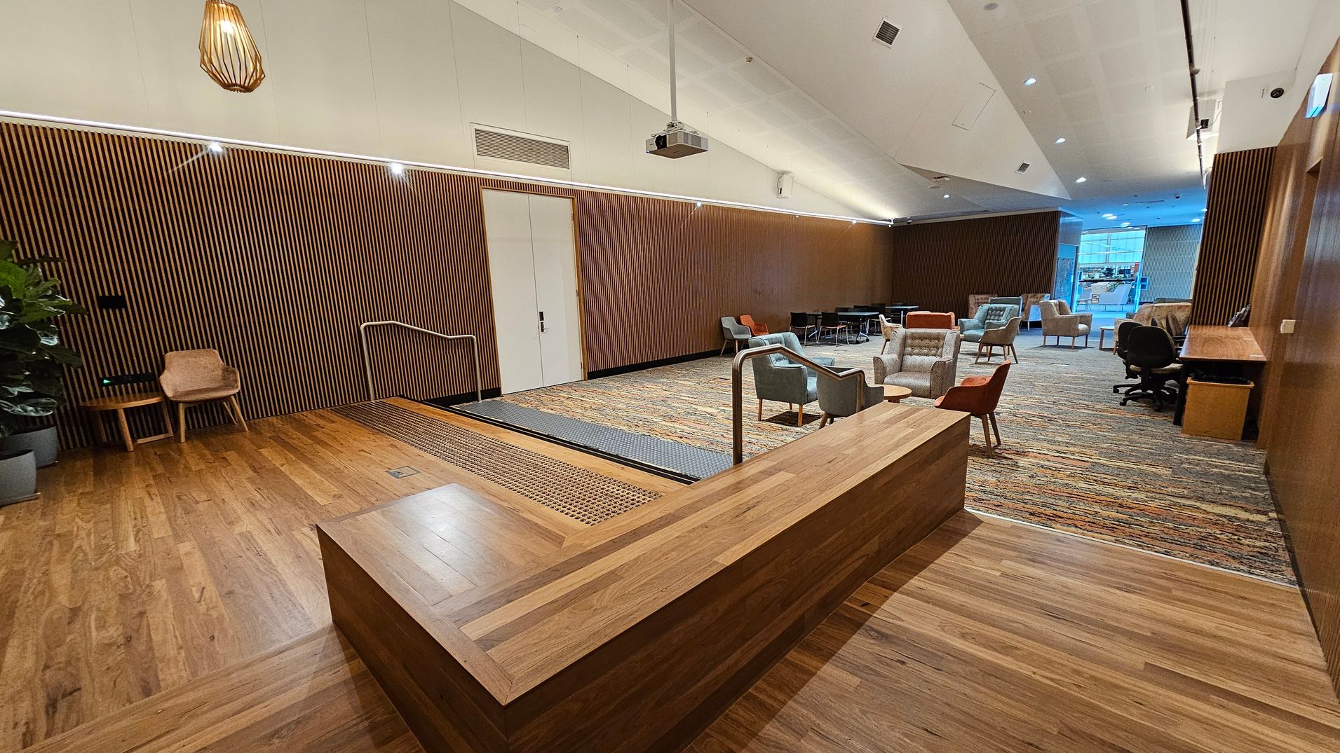 A modern interior space with a wooden floor and a slatted wooden wall on the left. A small wooden chair and table are near the wall. A ramp with metal railings leads to a carpeted area with various chairs and tables. A wooden counter is in the foreground. The ceiling is white with recessed lighting and a hanging light fixture. In the background, large windows allow natural light to enter, and there is a desk with an office chair on the right. A modern interior space with a wooden floor and a slatted wooden wall on the left. A small wooden chair and table are near the wall. A ramp with metal railings leads to a carpeted area with various chairs and tables. A wooden counter is in the foreground. The ceiling is white with recessed lighting and a hanging light fixture. In the background, large windows allow natural light to enter, and there is a desk with an office chair on the right.