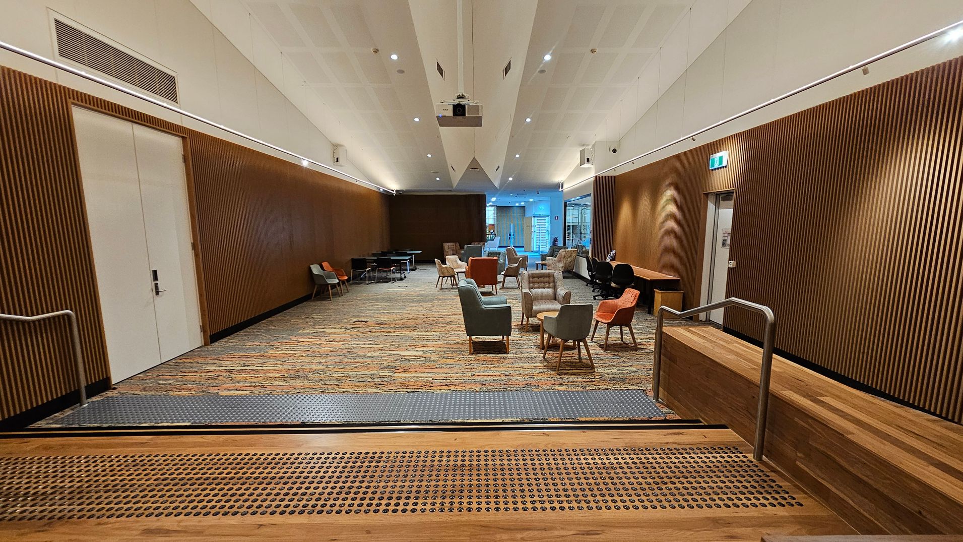 A spacious room with a patterned carpet floor and wooden panelled walls. In the centre, there are various chairs and small tables arranged in clusters. On the left, a large white door is set into the wall. On the right, a wooden desk and more seating are visible. The ceiling is white with recessed lighting and a projector. At the foreground, there are steps with a metal handrail leading down into the room. A spacious room with a patterned carpet floor and wooden panelled walls. In the centre, there are various chairs and small tables arranged in clusters. On the left, a large white door is set into the wall. On the right, a wooden desk and more seating are visible. The ceiling is white with recessed lighting and a projector. At the foreground, there are steps with a metal handrail leading down into the room.