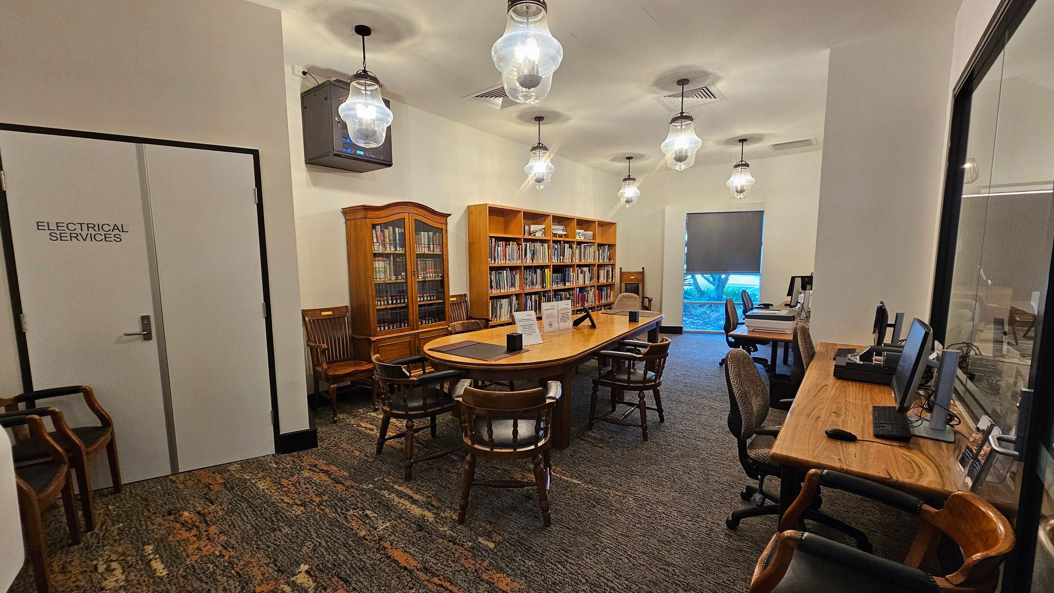 A library room with a carpeted floor. On the left, a door labelled "Electrical Services" and several wooden chairs. In the centre, a long wooden table surrounded by chairs, with bookshelves filled with books behind it. On the right, a wooden desk with computers and office chairs. The ceiling features several hanging light fixtures. A window with a partially drawn blind is in the background. A library room with a carpeted floor. On the left, a door labelled "Electrical Services" and several wooden chairs. In the centre, a long wooden table surrounded by chairs, with bookshelves filled with books behind it. On the right, a wooden desk with computers and office chairs. The ceiling features several hanging light fixtures. A window with a partially drawn blind is in the background.