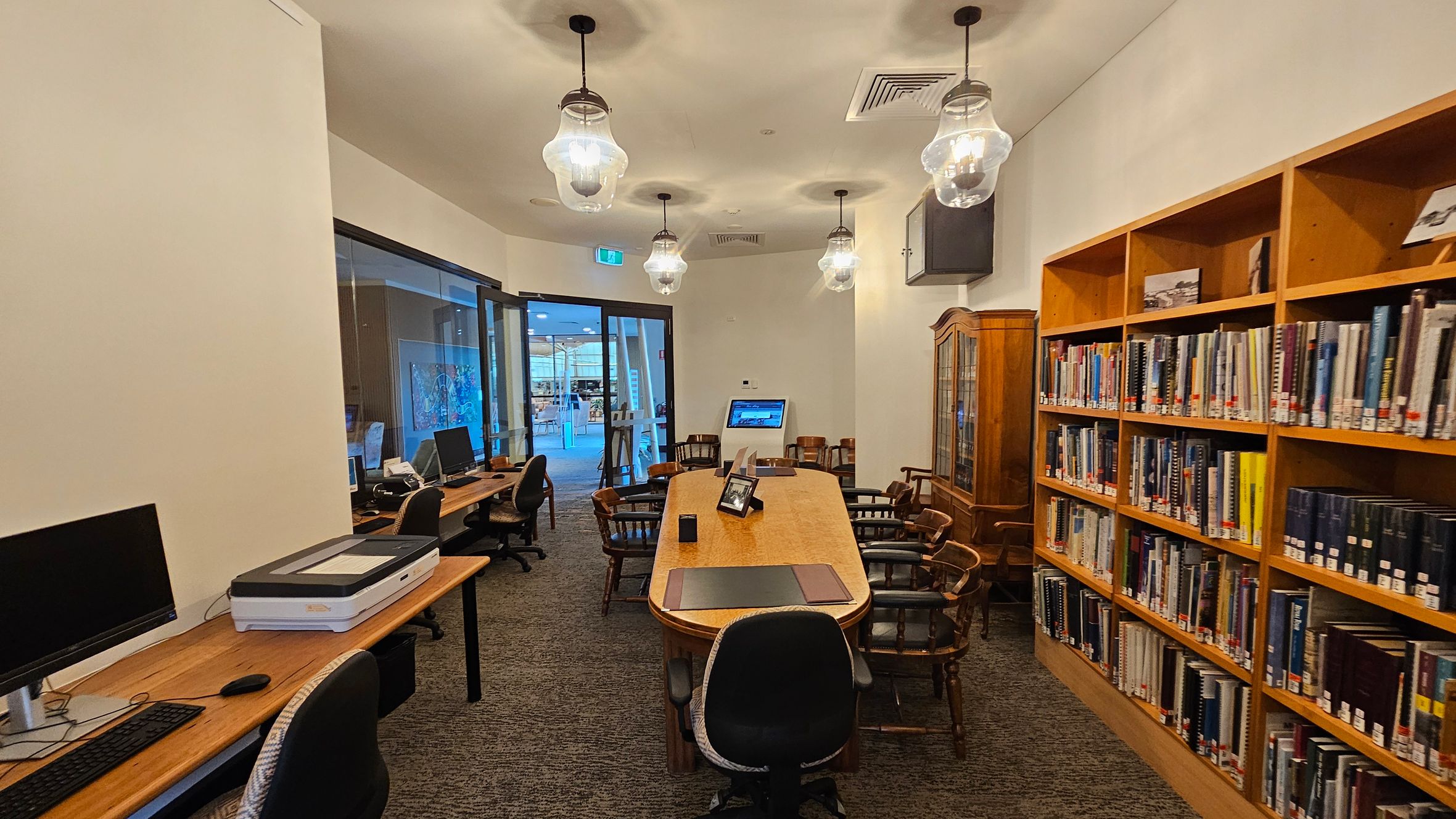 A library room with a long wooden table in the centre surrounded by wooden chairs. On the right, there are bookshelves filled with books. On the left, a row of desks with computers and a scanner. The floor is carpeted, and the ceiling has several hanging light fixtures. At the back, a glass door leads to another area with visible artwork and more seating. A library room with a long wooden table in the centre surrounded by wooden chairs. On the right, there are bookshelves filled with books. On the left, a row of desks with computers and a scanner. The floor is carpeted, and the ceiling has several hanging light fixtures. At the back, a glass door leads to another area with visible artwork and more seating.