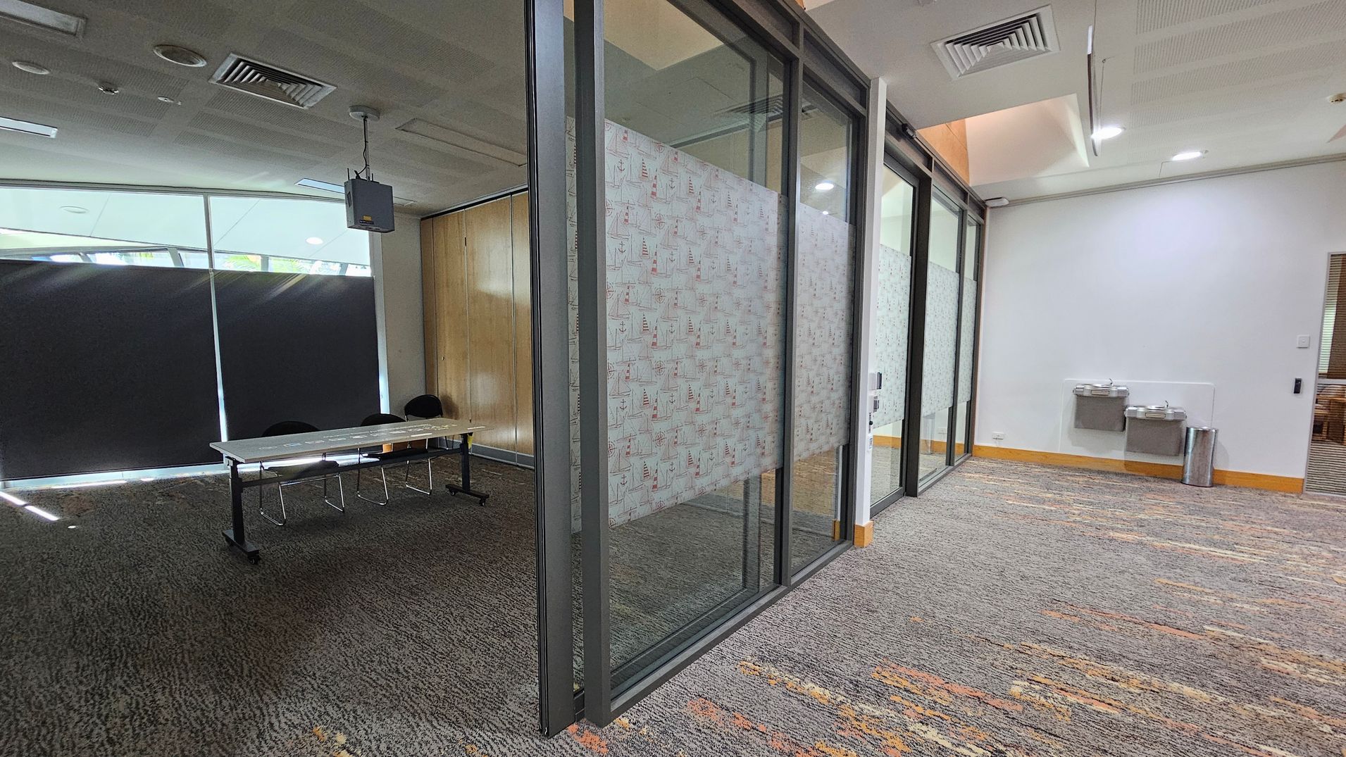 On the left, there is a room with a long table and several chairs, partially visible through a glass partition. The glass has a patterned design. The floor is carpeted with a mix of grey and brown tones. On the right, there are two water fountains mounted on a white wall, with a silver bin nearby. The ceiling has recessed lighting and air vents. On the left, there is a room with a long table and several chairs, partially visible through a glass partition. The glass has a patterned design. The floor is carpeted with a mix of grey and brown tones. On the right, there are two water fountains mounted on a white wall, with a silver bin nearby. The ceiling has recessed lighting and air vents.