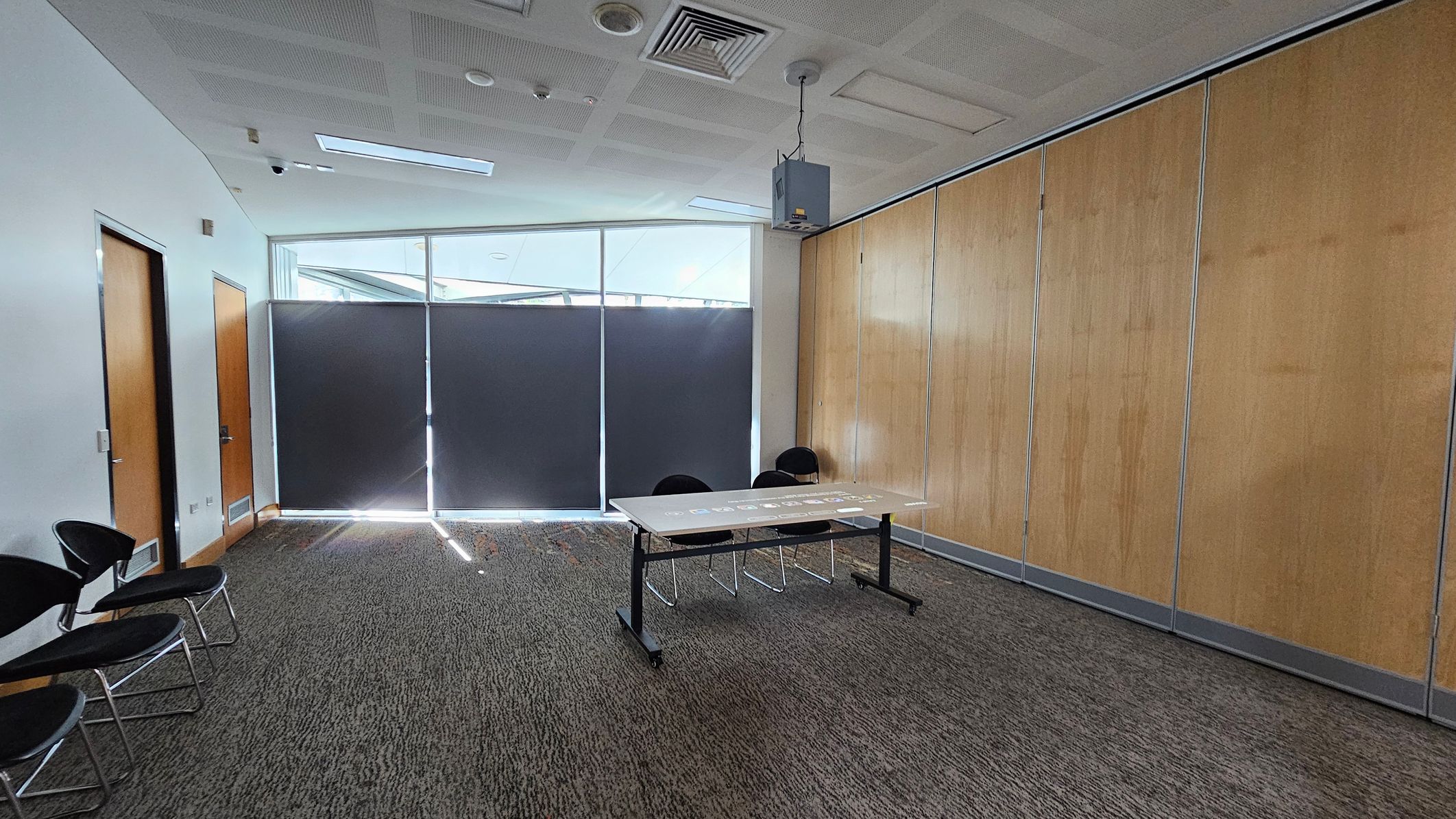 A room with a grey carpeted floor and a white ceiling featuring recessed lighting and vents. On the left, there are two wooden doors and a row of black chairs with metal legs. The centre has a table with two chairs behind it. The right side has a wall with light wooden panels. The back wall has large windows covered with dark blinds, allowing some light to filter through. A projector is suspended from the ceiling. A room with a grey carpeted floor and a white ceiling featuring recessed lighting and vents. On the left, there are two wooden doors and a row of black chairs with metal legs. The centre has a table with two chairs behind it. The right side has a wall with light wooden panels. The back wall has large windows covered with dark blinds, allowing some light to filter through. A projector is suspended from the ceiling.