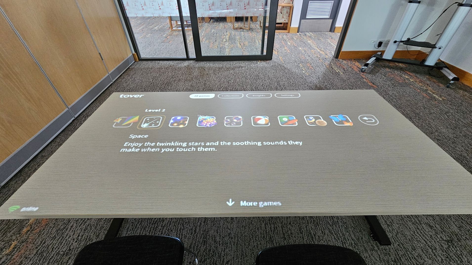 A digital interface is projected onto a table, displaying a game menu with icons and text. The text reads, "Enjoy the twinkling stars and the soothing sounds they make when you touch them." In the background, there is a glass partition with a view into another room, which has a carpeted floor and a wooden chair. The floor is carpeted with a pattern, and there are wooden panels on the left wall. Two black chairs are partially visible in the foreground. A digital interface is projected onto a table, displaying a game menu with icons and text. The text reads, "Enjoy the twinkling stars and the soothing sounds they make when you touch them." In the background, there is a glass partition with a view into another room, which has a carpeted floor and a wooden chair. The floor is carpeted with a pattern, and there are wooden panels on the left wall. Two black chairs are partially visible in the foreground.