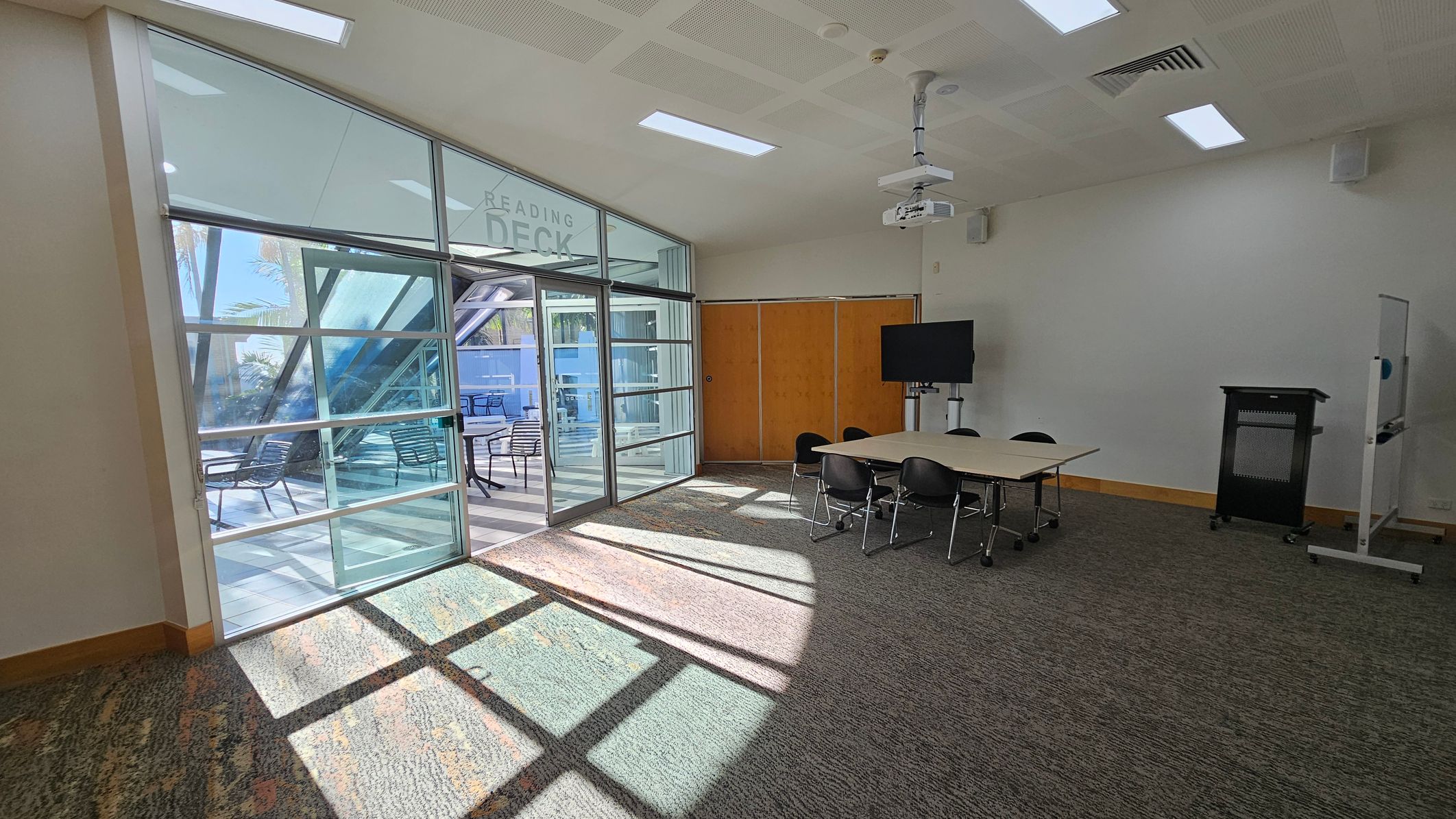 A room with a carpeted floor and a ceiling featuring recessed lighting and a projector. On the right, there's a table with several chairs, a TV on a stand, and a lectern. A whiteboard is also visible. On the left, large glass doors lead to an outdoor area with tables and chairs, labelled "Reading Deck." Sunlight streams through the glass, casting shadows on the floor. A room with a carpeted floor and a ceiling featuring recessed lighting and a projector. On the right, there's a table with several chairs, a TV on a stand, and a lectern. A whiteboard is also visible. On the left, large glass doors lead to an outdoor area with tables and chairs, labelled "Reading Deck." Sunlight streams through the glass, casting shadows on the floor.