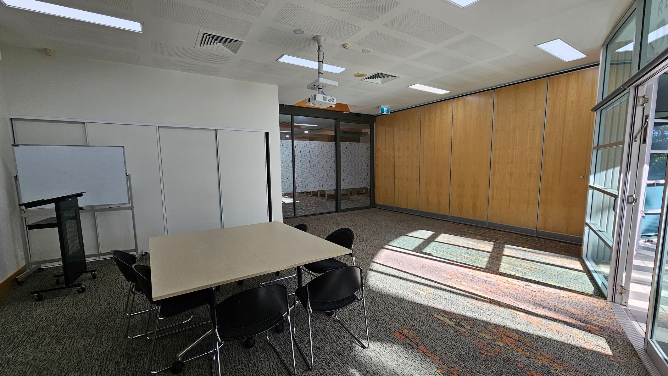 A conference room with a light-coloured table surrounded by black chairs on the left. A whiteboard and a lectern are positioned against the left wall. The right side features large wooden panels and glass doors, allowing natural light to stream in, casting shadows on the carpeted floor. The ceiling has recessed lighting and a mounted projector. A conference room with a light-coloured table surrounded by black chairs on the left. A whiteboard and a lectern are positioned against the left wall. The right side features large wooden panels and glass doors, allowing natural light to stream in, casting shadows on the carpeted floor. The ceiling has recessed lighting and a mounted projector.