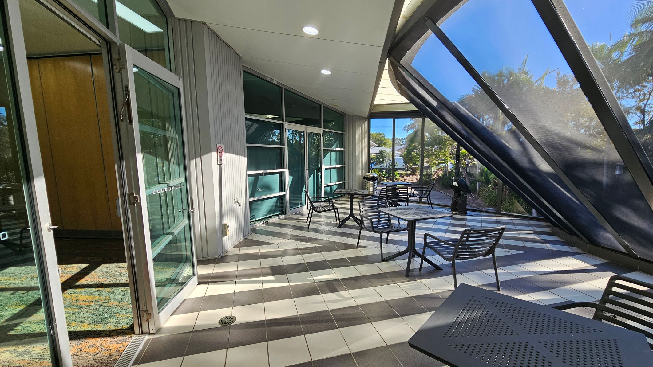A modern indoor space with a slanted glass wall on the right, allowing natural light to flood in. The floor is tiled in a checkered pattern of grey and white. On the left, there's a glass door leading to an interior area with a patterned carpet. Several black metal tables and chairs are arranged throughout the space. In the background, greenery is visible through the glass, and a small sculpture is near the window. The ceiling is smooth with recessed lighting. A modern indoor space with a slanted glass wall on the right, allowing natural light to flood in. The floor is tiled in a checkered pattern of grey and white. On the left, there's a glass door leading to an interior area with a patterned carpet. Several black metal tables and chairs are arranged throughout the space. In the background, greenery is visible through the glass, and a small sculpture is near the window. The ceiling is smooth with recessed lighting.