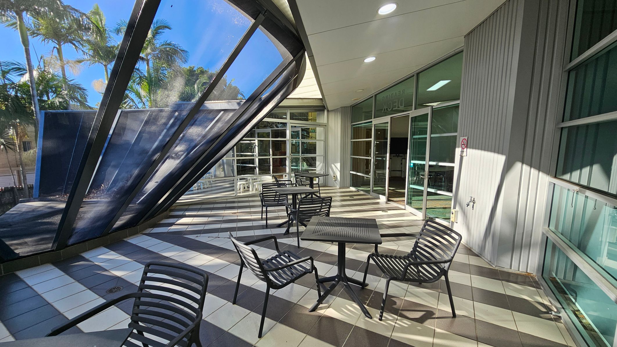 Outdoor seating area with black metal tables and chairs on a tiled floor featuring a striped pattern. On the left, a large, slanted glass structure with metal frames provides partial shade. In the background, tall palm trees are visible. The right side has a building with large glass doors and windows, and a small wall-mounted tap. The ceiling is white with recessed lighting. Outdoor seating area with black metal tables and chairs on a tiled floor featuring a striped pattern. On the left, a large, slanted glass structure with metal frames provides partial shade. In the background, tall palm trees are visible. The right side has a building with large glass doors and windows, and a small wall-mounted tap. The ceiling is white with recessed lighting.