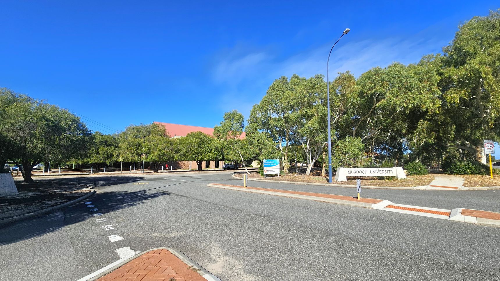 A road curves gently to the left, bordered by trees on both sides. On the right, a sign reads "Murdoch University" near a lamppost. A smaller sign and a speed limit sign are also visible. The left side features more trees and a glimpse of a red-roofed building in the background. The road surface is grey, with a brick-patterned section in the foreground.