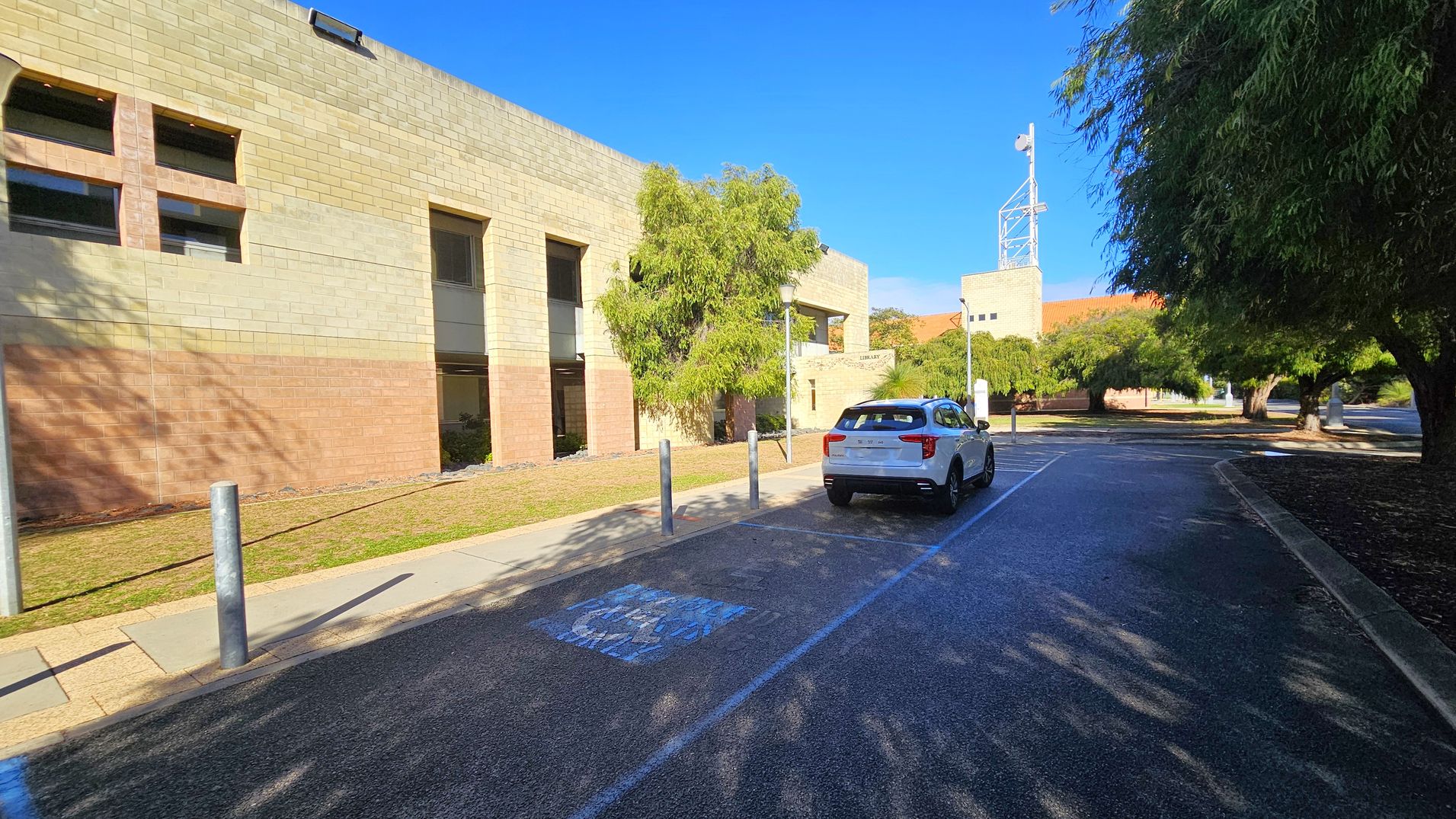A paved road with a white car parked on the right side. On the left, there's a large beige brick building with rectangular windows and a small tree in front. The road has a marked parking space and is lined with bollards. Trees with dense foliage are on the right, casting shadows on the road. A tall structure with antennas is visible in the background. The area is well-lit, suggesting a sunny day.