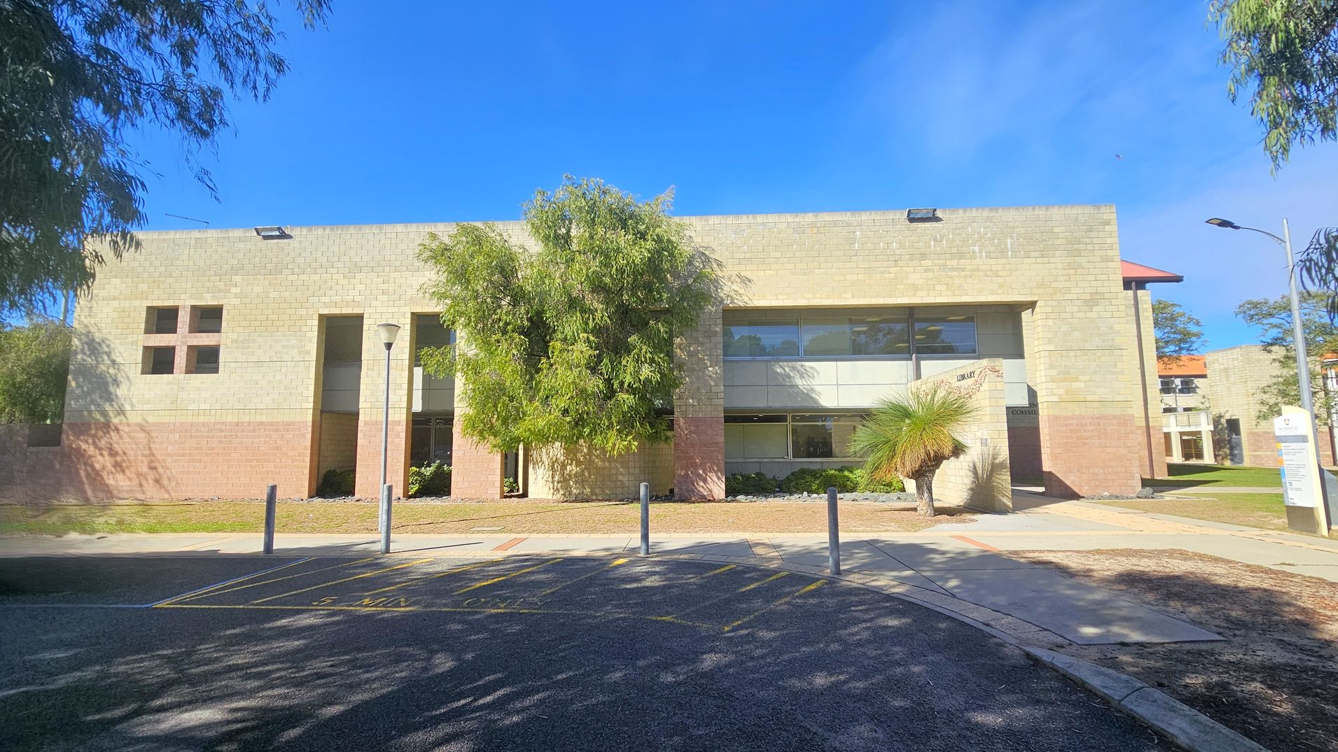 A two-storey building with a beige and brown brick facade. Large windows are on the upper and lower levels. A tree is in front of the building, partially obscuring the view. A small sign near the entrance reads "Library." The foreground features a paved area with parking lines and bollards. A lamp post is visible on the left, and another building is partially visible on the right. Trees frame the scene on both sides.