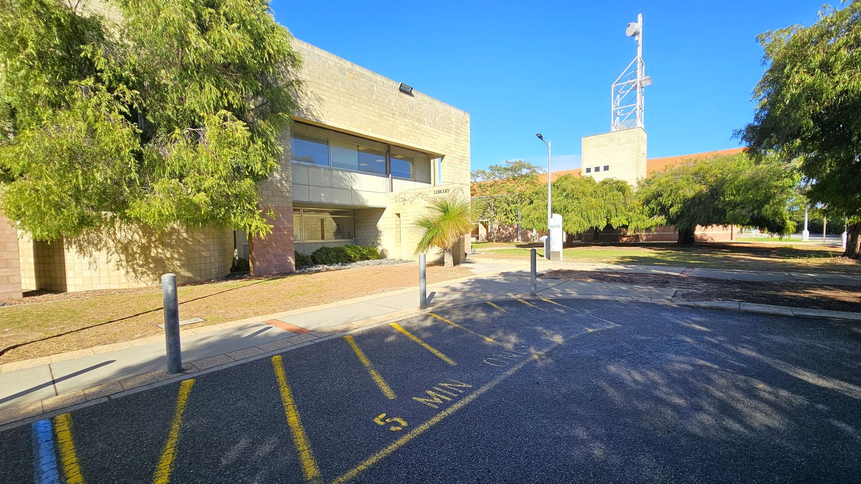 A beige brick building with large windows is on the left, partially obscured by lush green trees. The word "Library" is visible on the building. In the foreground, a paved area with yellow markings reads "5 MIN." Several bollards line the edge of the pavement. On the right, more trees provide shade, and a tall structure with a metal framework is visible in the background. The ground is a mix of grass and pavement.