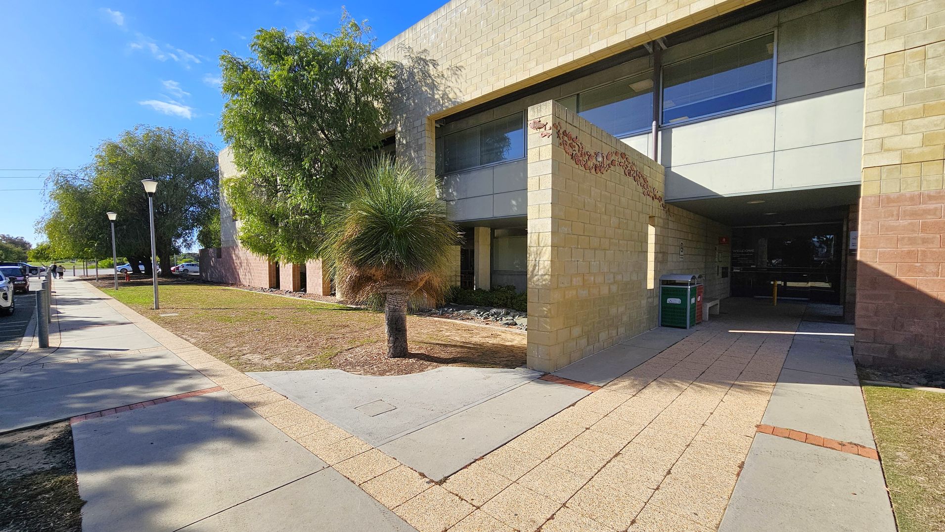 A modern building with light-coloured brick walls and large windows on the right. The entrance is recessed, with a pathway leading to it. A green bin is near the entrance. On the left, a paved walkway runs alongside the building, lined with street lamps and trees. Several parked cars are visible along the street. The ground is a mix of grass and concrete, with a small palm-like tree near the building.